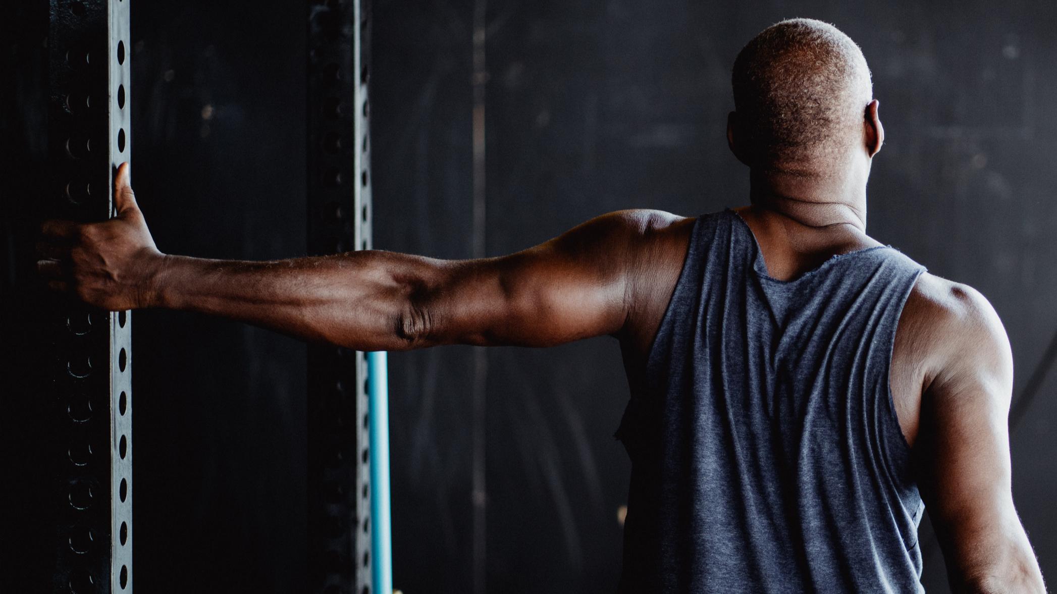 A muscular man with a shaved head wearing a sleeveless dark gray shirt stretches his arm against a black metal wall or rack in a gym setting.