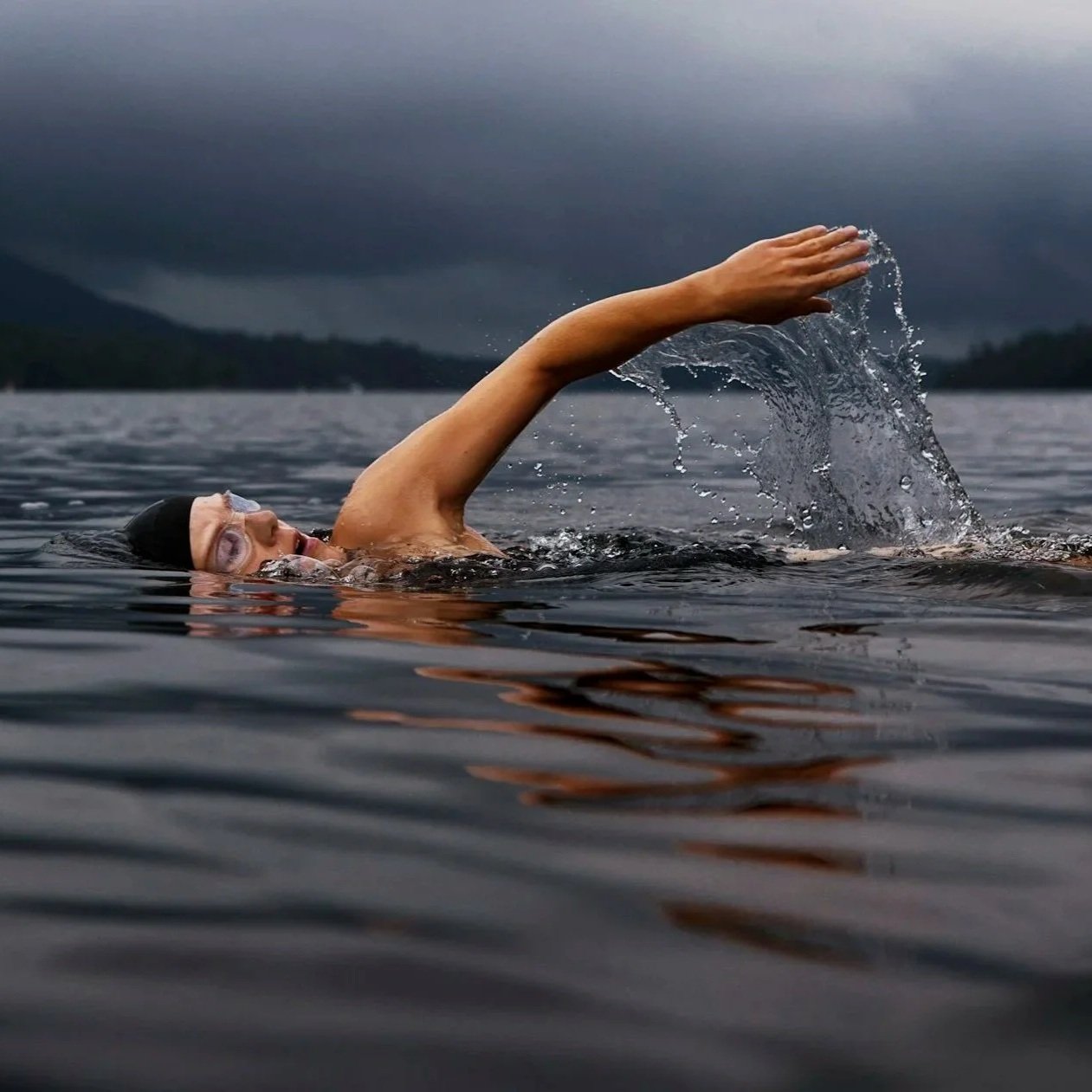 A person swimming in open water, wearing goggles and a black swim cap, with a cloudy sky and distant mountains in the background.