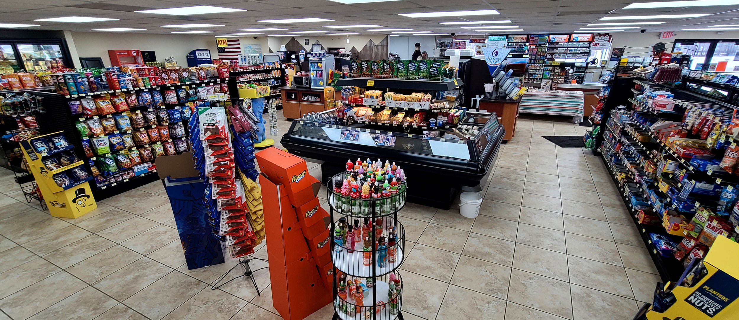 Inside the Fuel On convenience store in Auburndale, Wisconsin, showing clean, wide aisles, fully stocked product, a refrigerated air case, EZ-Tab vehicle registration machine, no-fee ATM, and a Red Bull cooler.