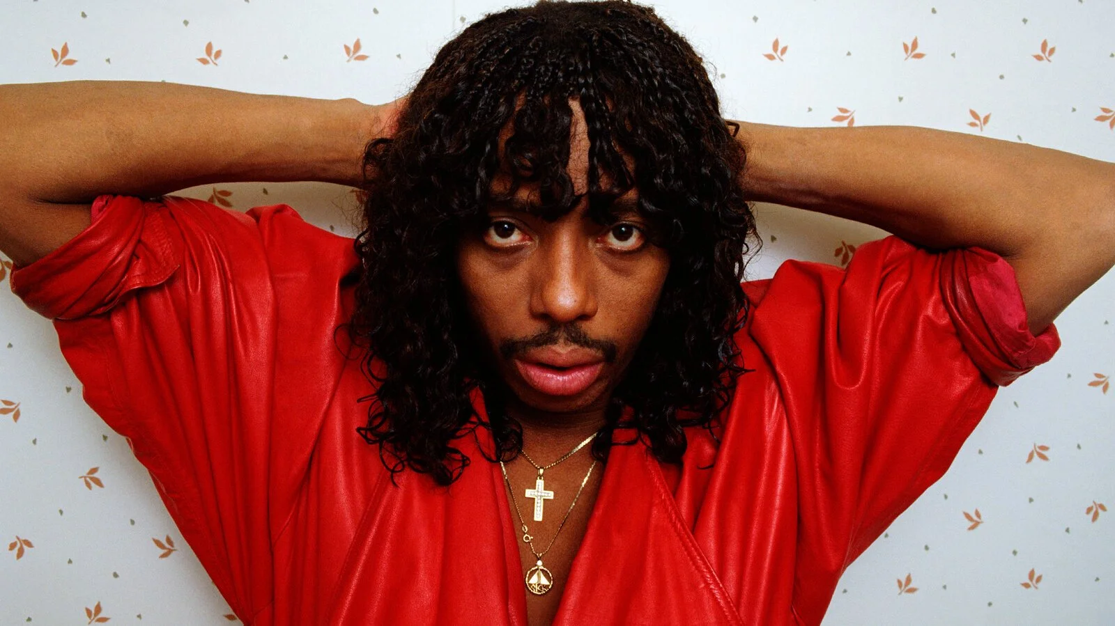A person with dark curly hair wearing a red leather jacket, gold jewelry including a cross necklace, posing with hands behind head against a white wall with small orange leaf patterns.