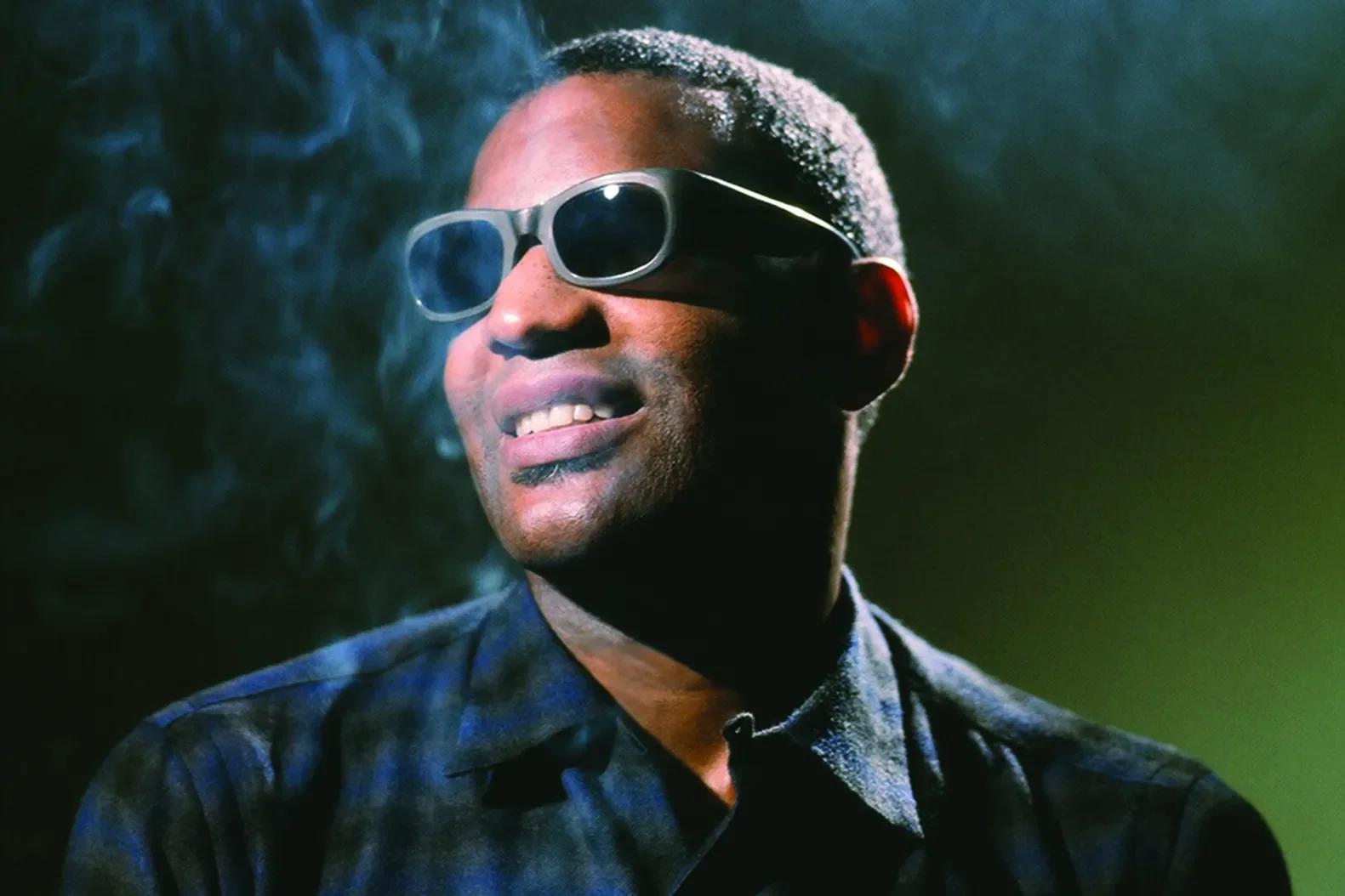 Man wearing black sunglasses, smiling, with a dark background and greenish lighting.