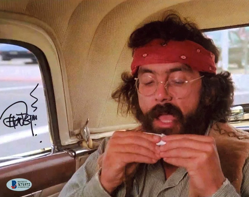 A man with curly hair, glasses, and a beard wearing a red bandana, sitting in a car with his eyes closed and hands under his chin, appearing to be sleeping or resting.