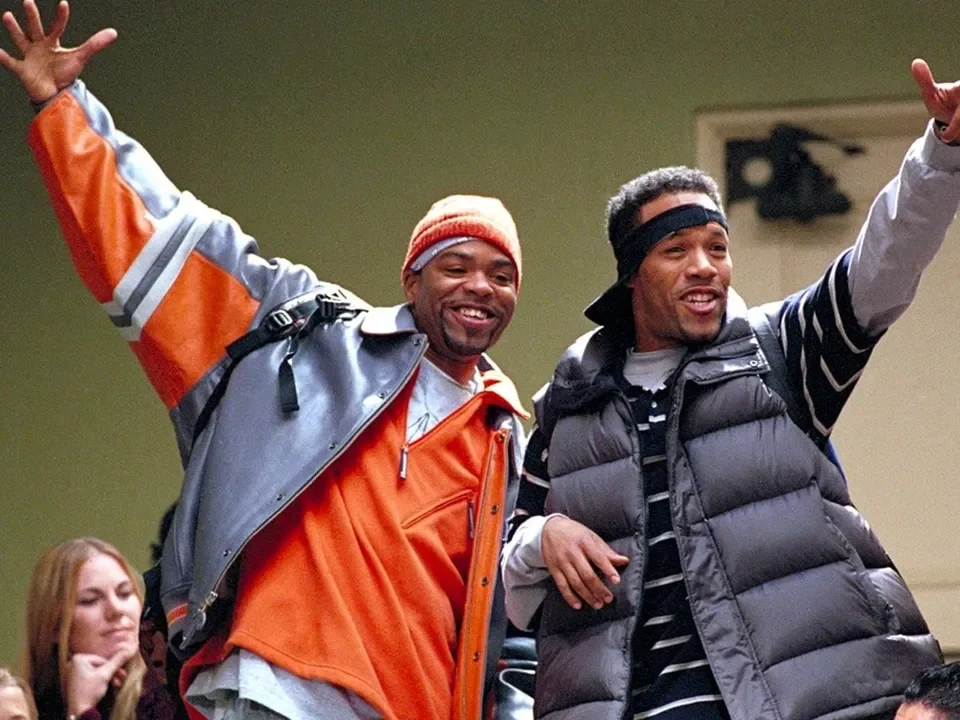 Two men smiling and gesturing with arms raised, wearing winter jackets and headbands, in an indoor setting, with a woman in the background watching them.