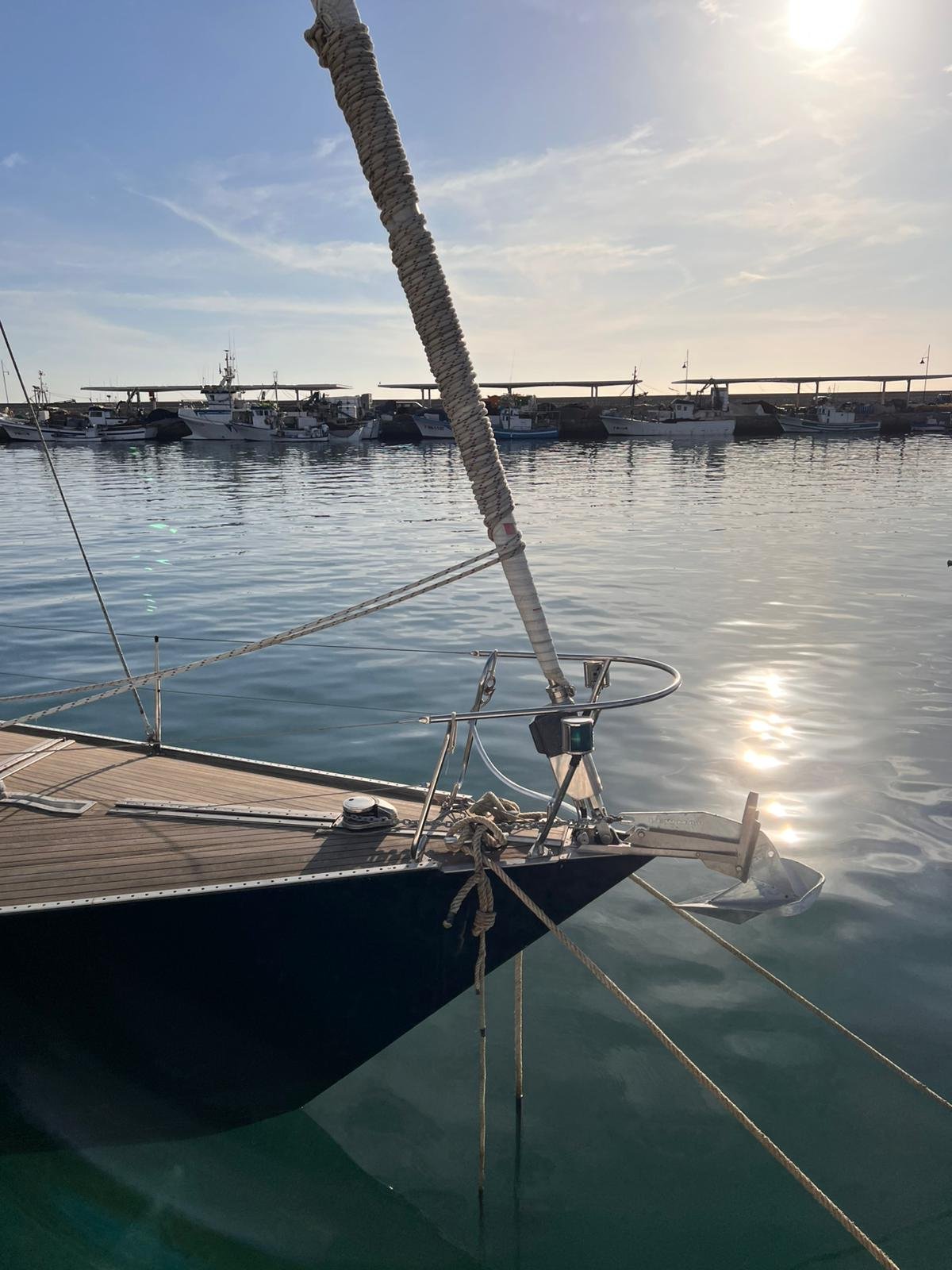 Close-up of the bow of a sailboat with a wooden deck, ropes, and a fender, docked in a harbor with many other boats and yachts in the background under a partly cloudy sky during sunset.
