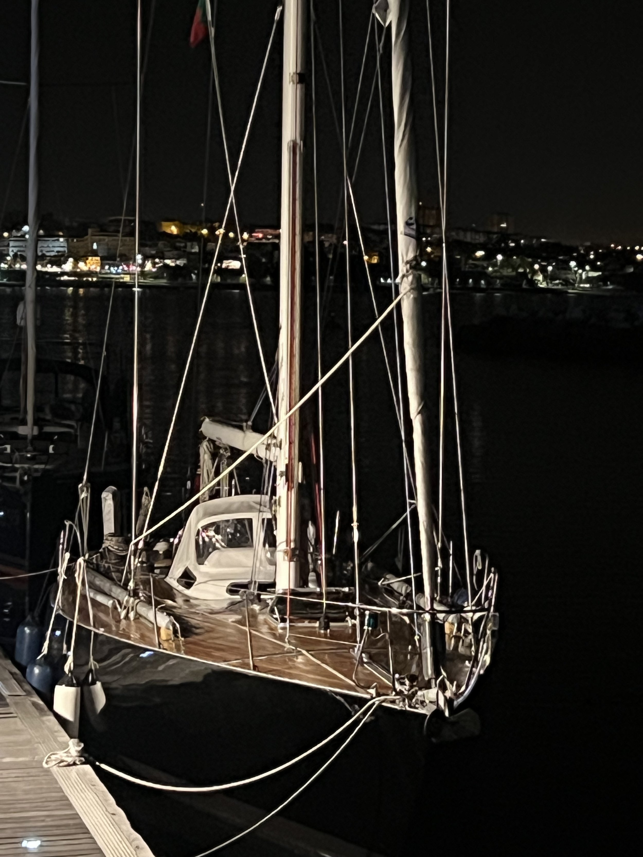 A sailboat docked at a marina at night with its mast and rigging visible, and city lights reflecting on the water in the background.