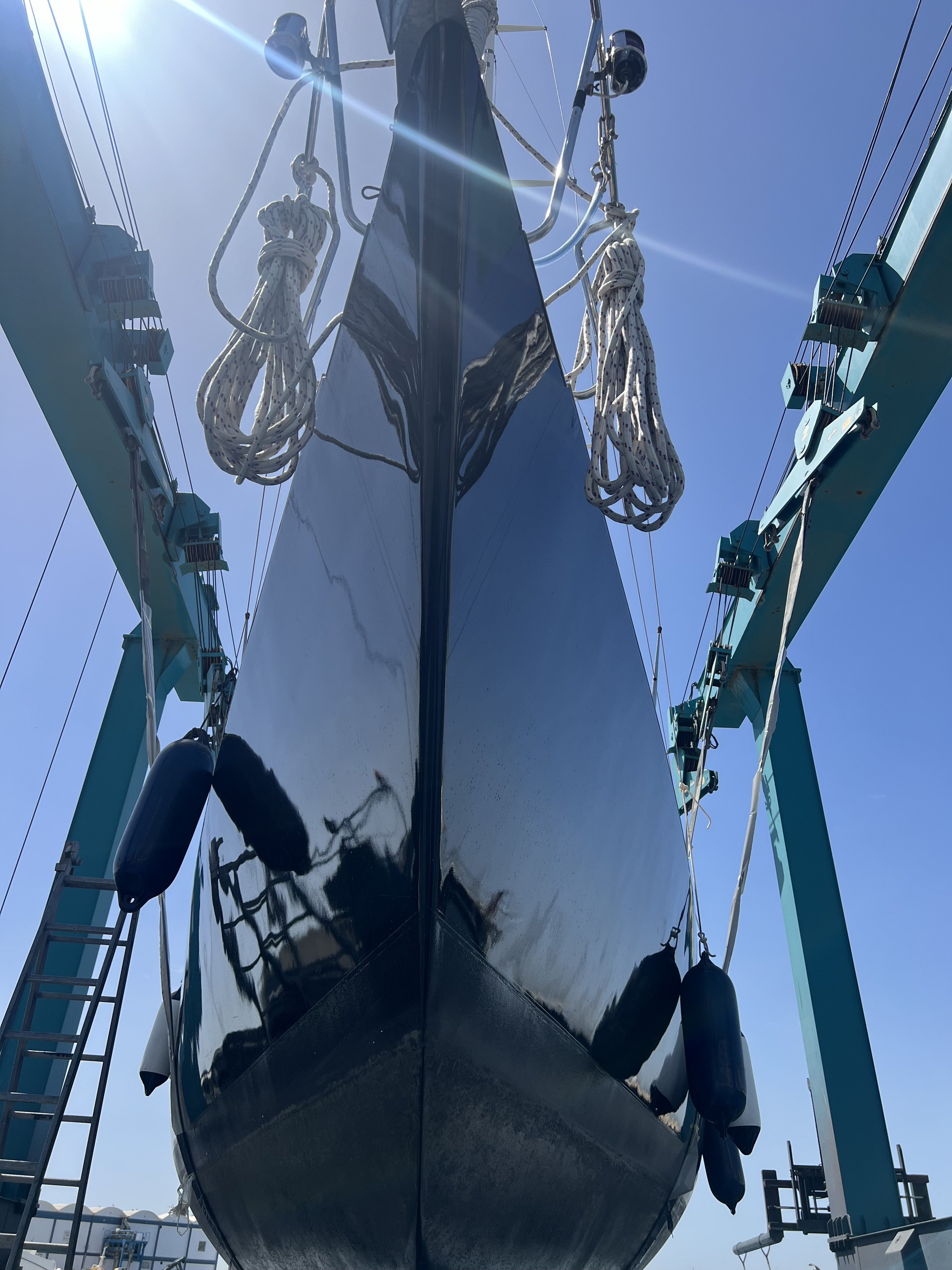 Looking up at a boat in a dry dock, with the hull reflected on its shiny black surface, surrounded by blue supports and ropes on a clear sunny day.