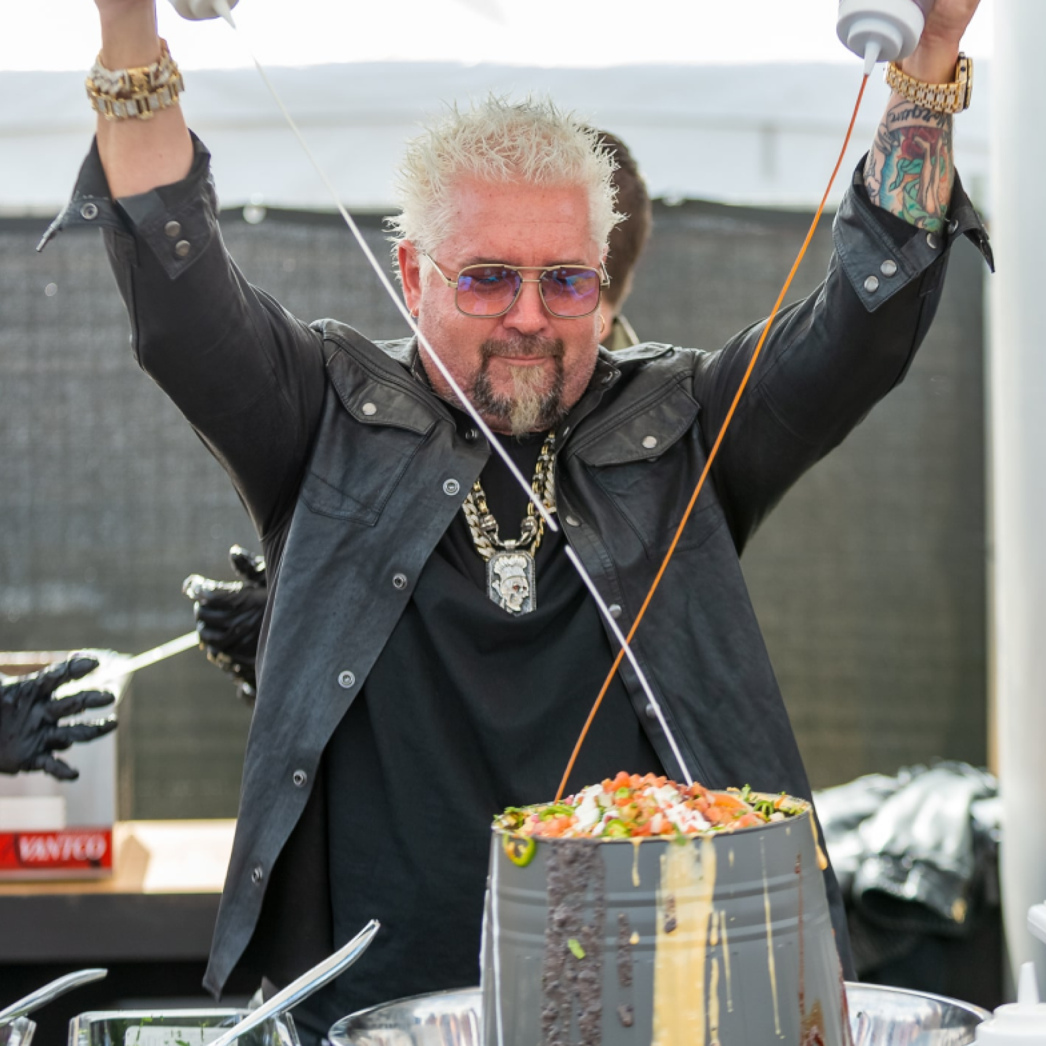 A man with bleached-blonde hair, sunglasses, a black leather jacket, and tattoos on his arms is holding a bottle of sauce over a large bowl of salad, smiling.
