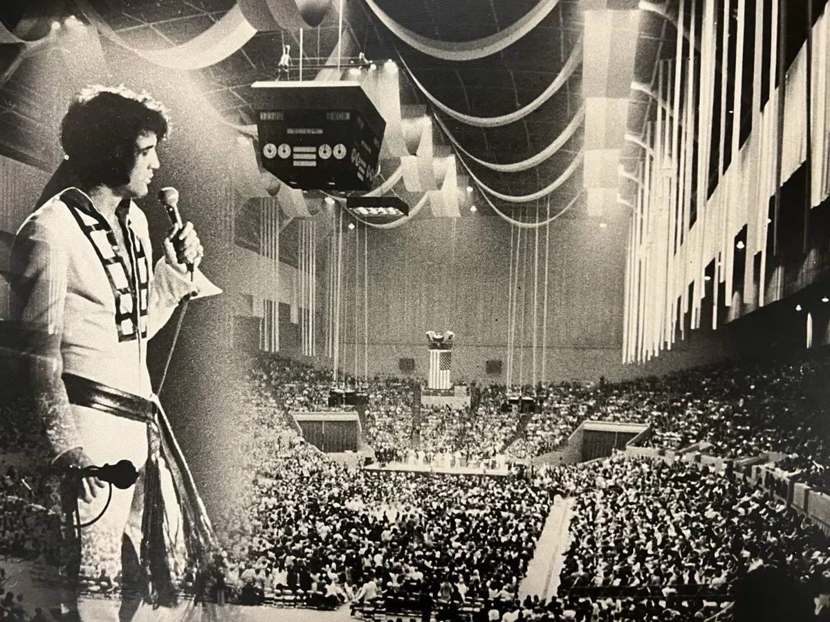 Elvis holding a microphone on stage in front of a large audience in an auditorium with high ceilings and decorative hanging banners.