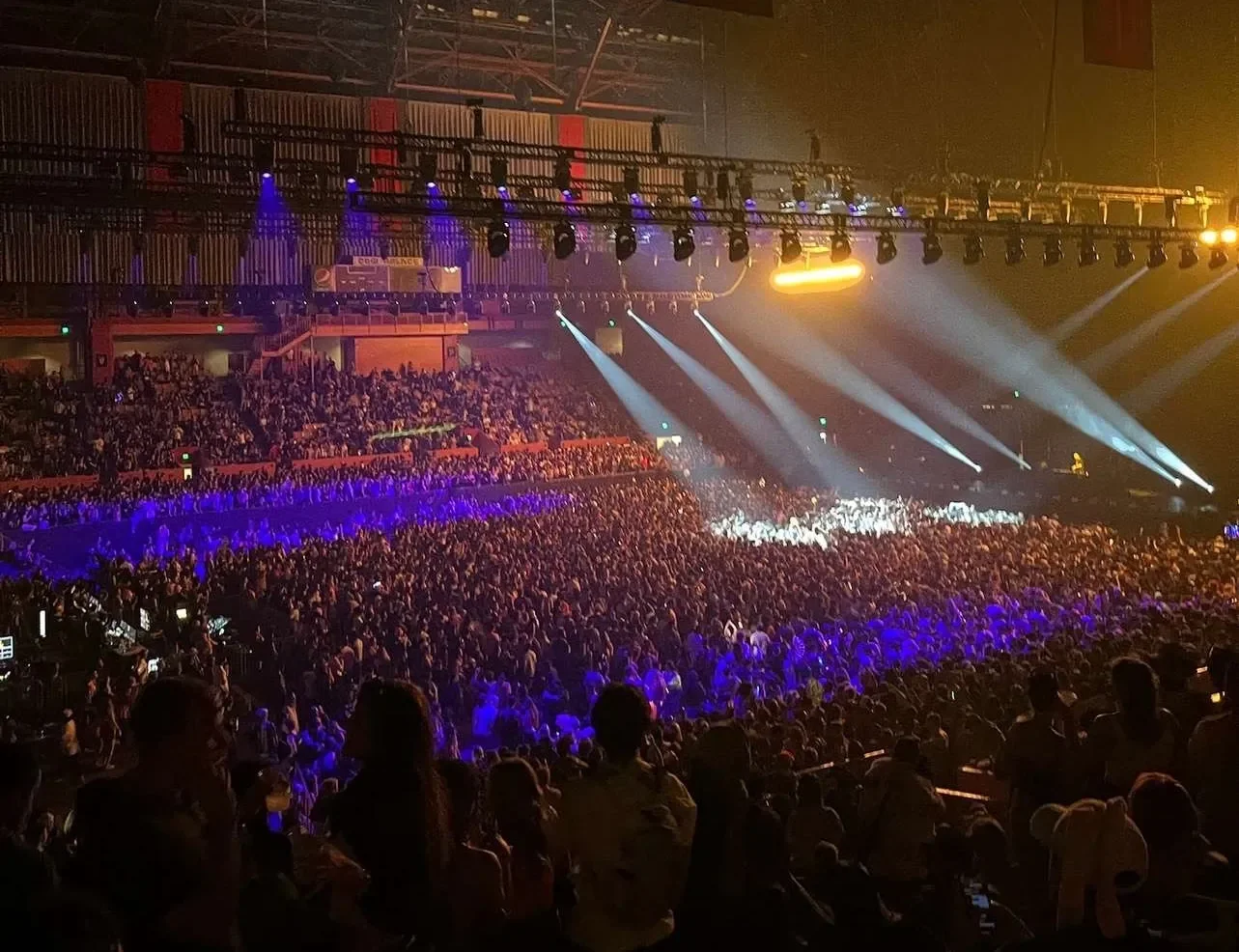 A large indoor concert with a massive crowd, stage lights, and colorful lighting effects in a sports arena.