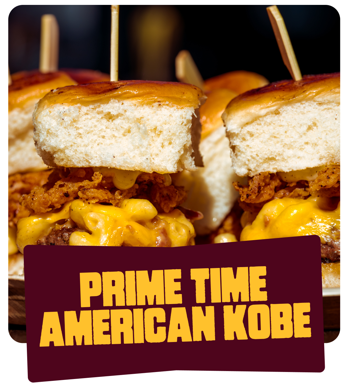 Close-up of cheeseburgers with buns, cheese, fried chicken, and toppings, with a sign reading 'Prime Time American Kobe' in the foreground.