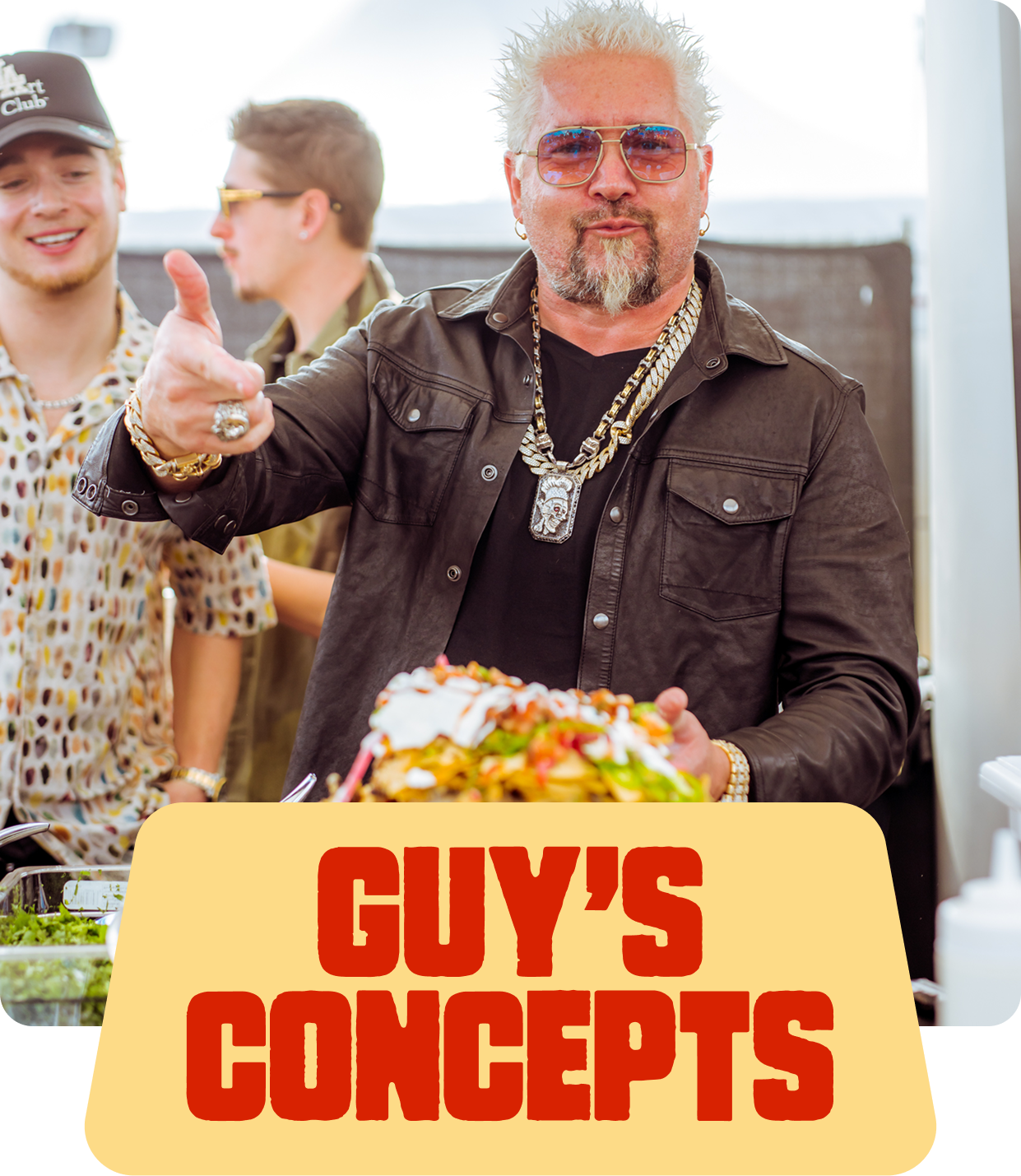 A man with white spiked hair, sunglasses, and jewelry giving a thumbs-up while holding food at a food stand, with two other young men in the background, and a sign that reads 'GUY'S CONCEPTS'.