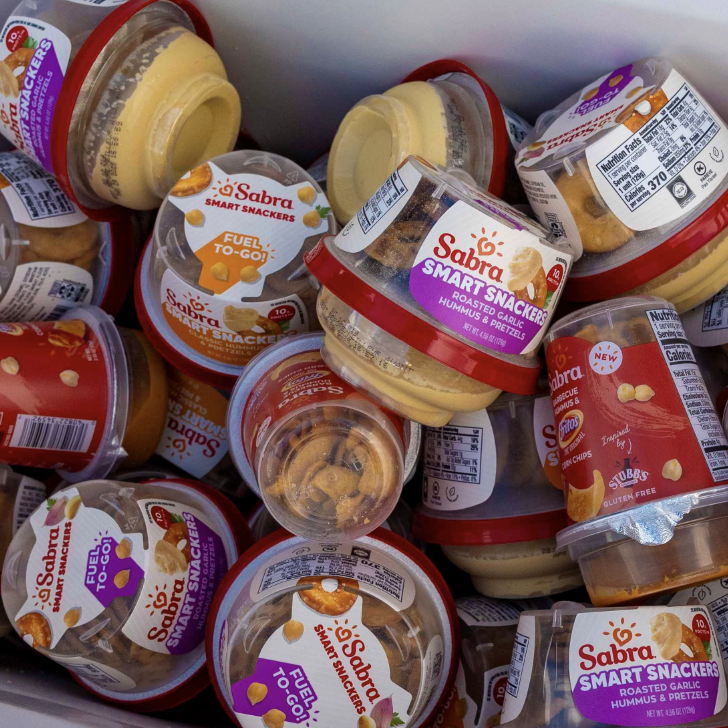 Multiple containers of Sabra smart snack hummus snack packs and chips, some opened, piled together.