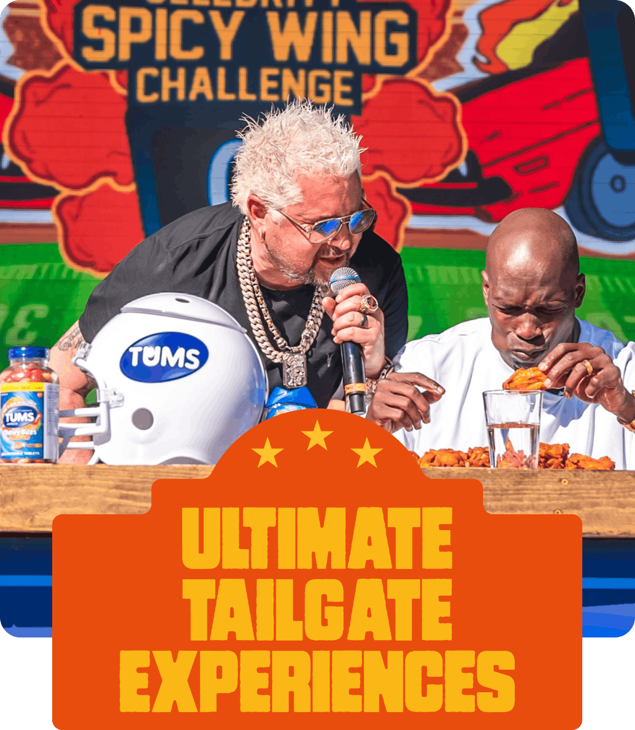 Two men sit at a table during an event called the "Spicy Wing Challenge". One man has spiky white hair, glasses, and is holding a microphone and a piece of chicken, while the other man is bald, appears to be grimacing as he eats spicy chicken wings. A TUMS helmet and antacid tablets are on the table, with a sign in the background reading "Ultimate Tailgate Experiences".