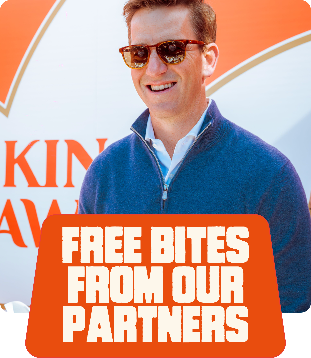 A man wearing sunglasses and a blue zip-up sweater standing in front of a sign that says "King something". There is a sign in front of him that reads "Free bites from our partners".
