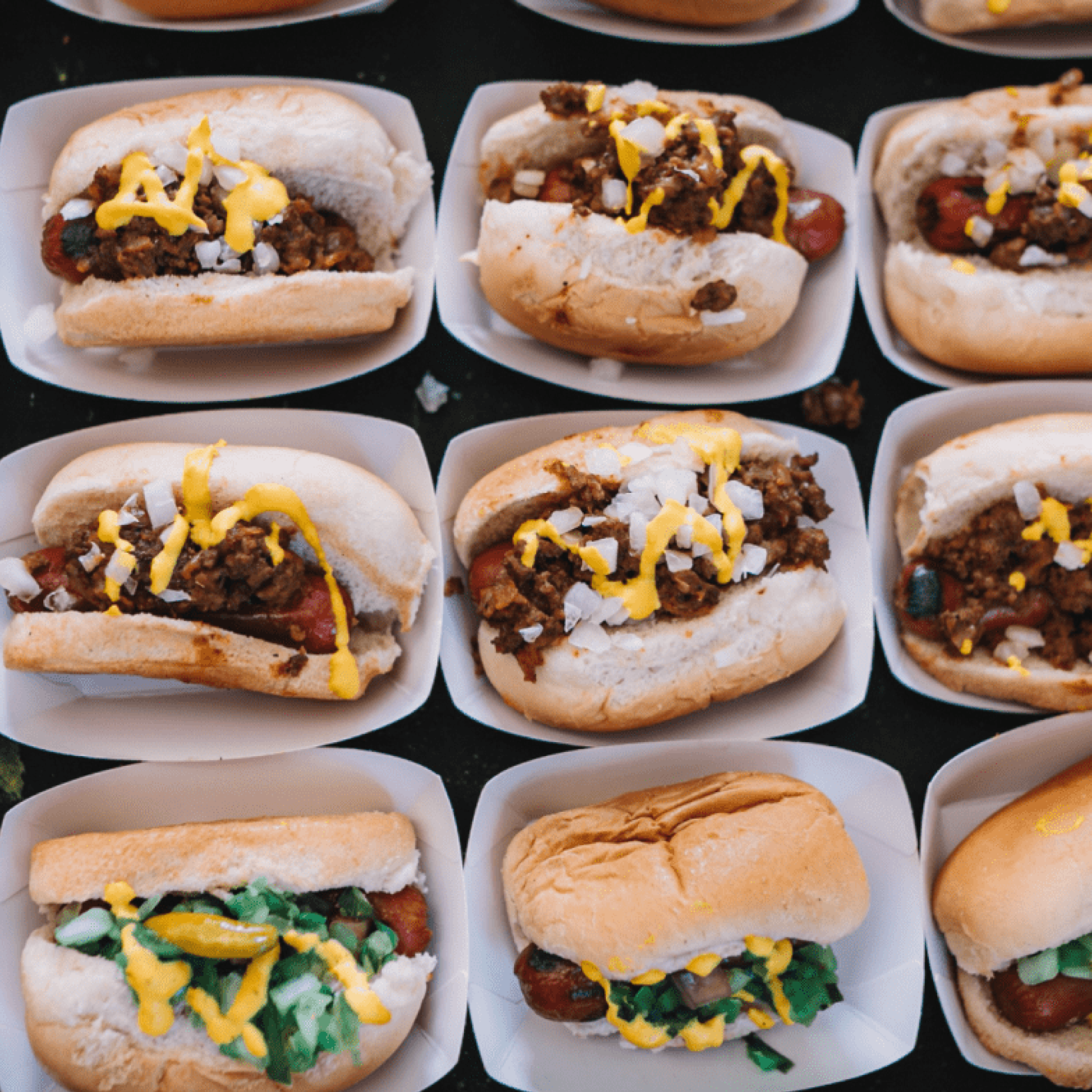 Multiple hot dogs in buns topped with mustard, chopped onions, and relish, arranged in paper trays.