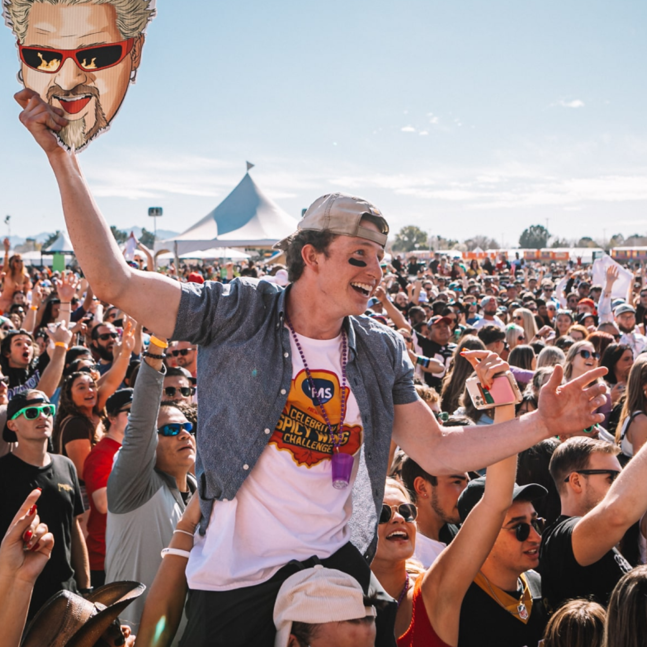 A young man at a crowded outdoor concert or festival, lifting a cardboard caricature of a person's face on a stick, wearing a baseball cap, sunglasses, and a white t-shirt, smiling and enjoying the event.