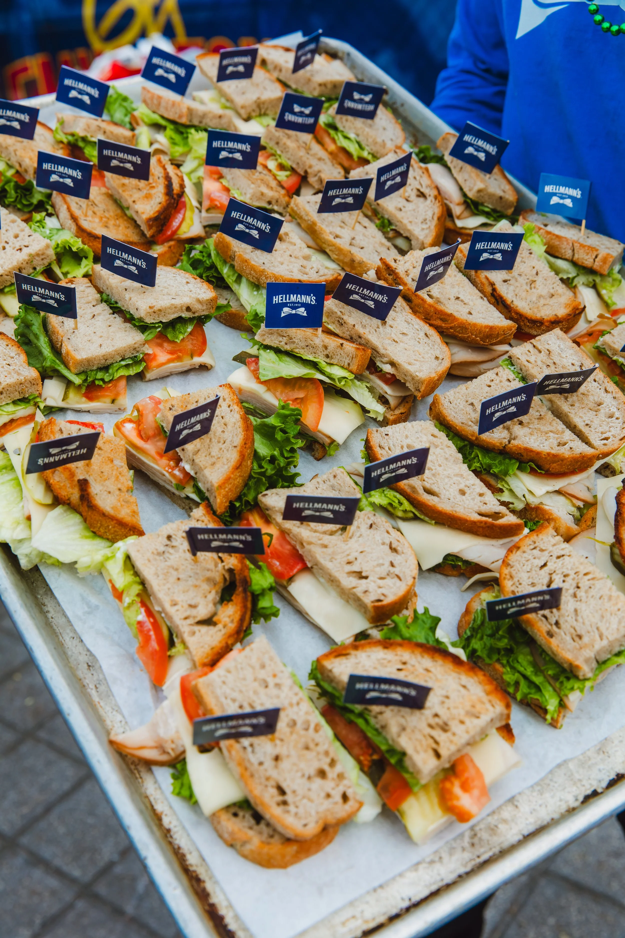 Assorted sandwiches with lettuce, tomato, cheese, and meat, arranged on a tray with small flag labels reading 'Hellmann's'.