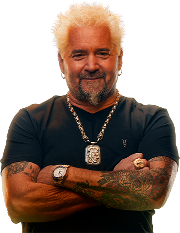 Man with platinum blonde hair and a beard, wearing a black t-shirt, multiple necklaces, a watch, and a ring, with tattoos on his arms, standing with his arms crossed.