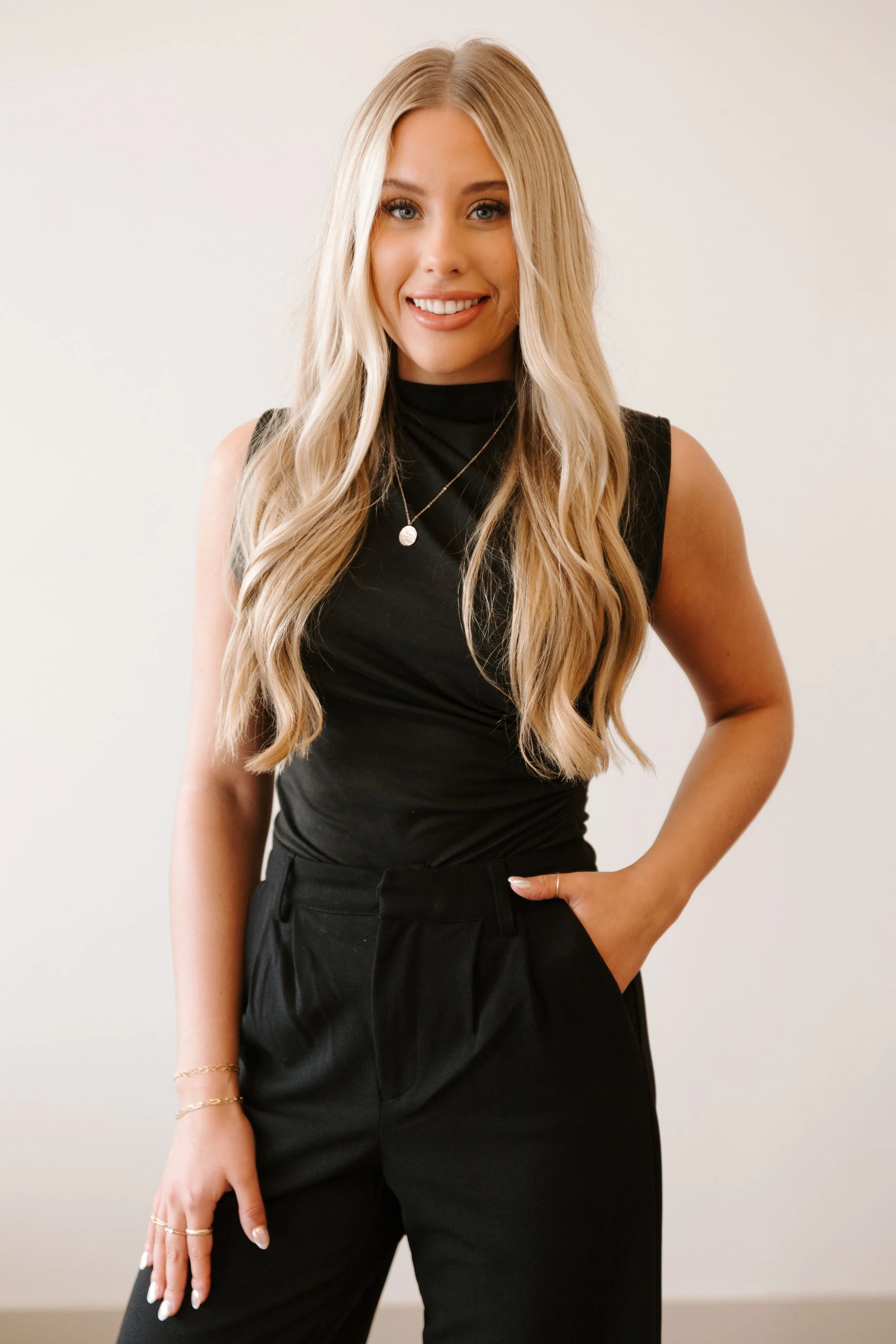 Woman with long blonde hair sitting on a stool by a window, wearing a black outfit and smiling.