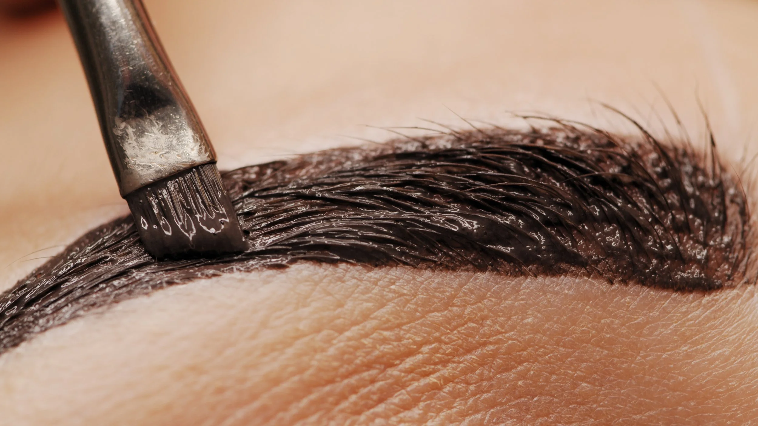 Close-up of a person's eyebrow being filled in with dark makeup using a brush.