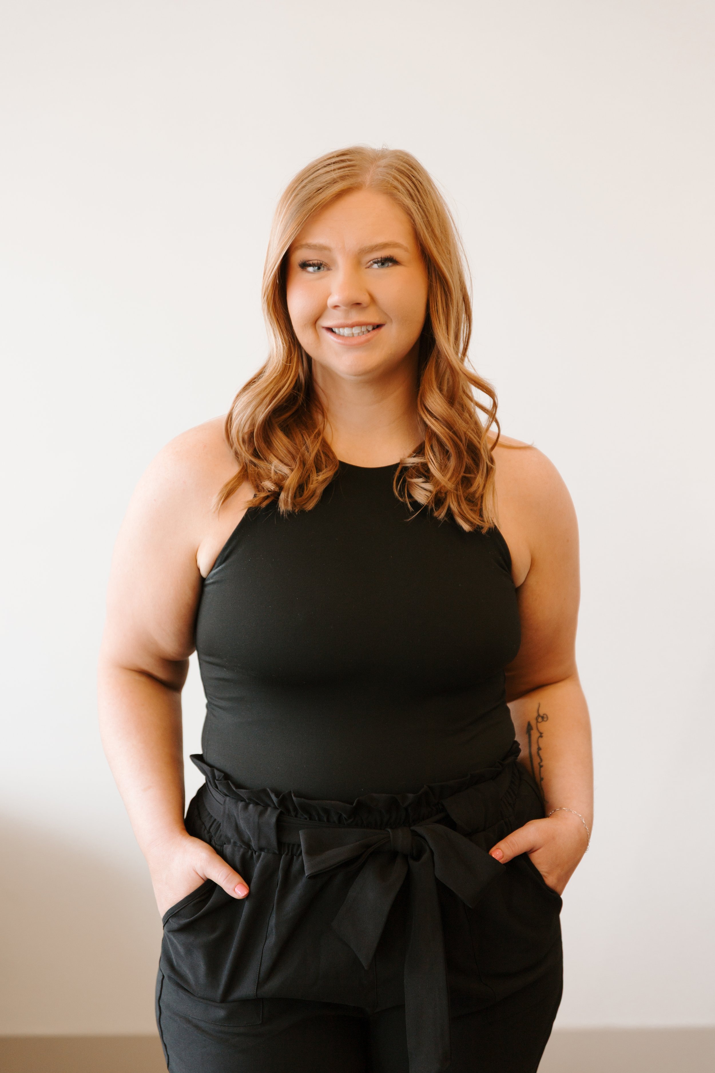 A woman with long, wavy red hair wearing a black sleeveless top and black pants with a bow, standing against a plain light-colored wall, smiling and looking at the camera.