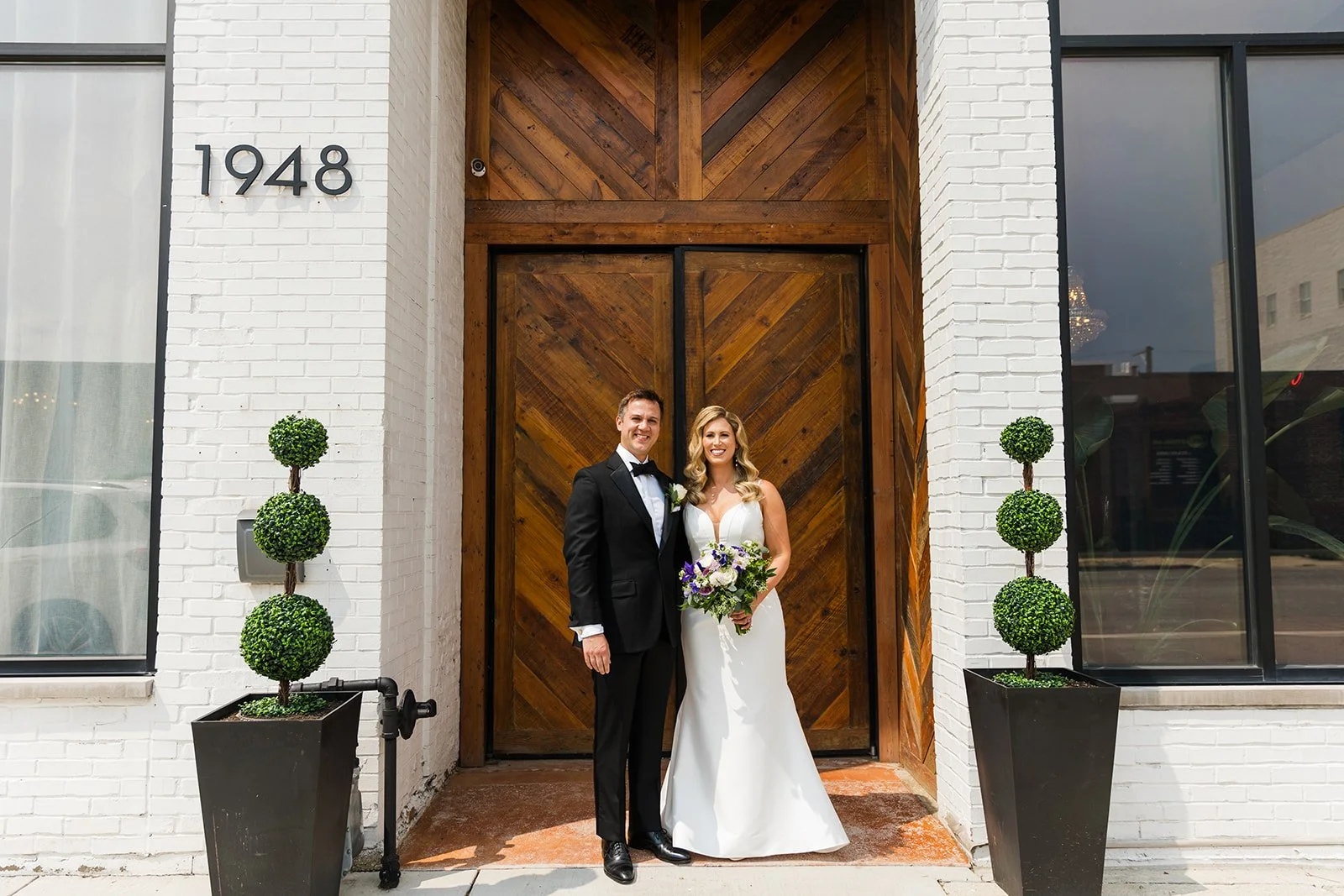 Wedding couple posing in front of wooden doors with white brick walls and greenery.