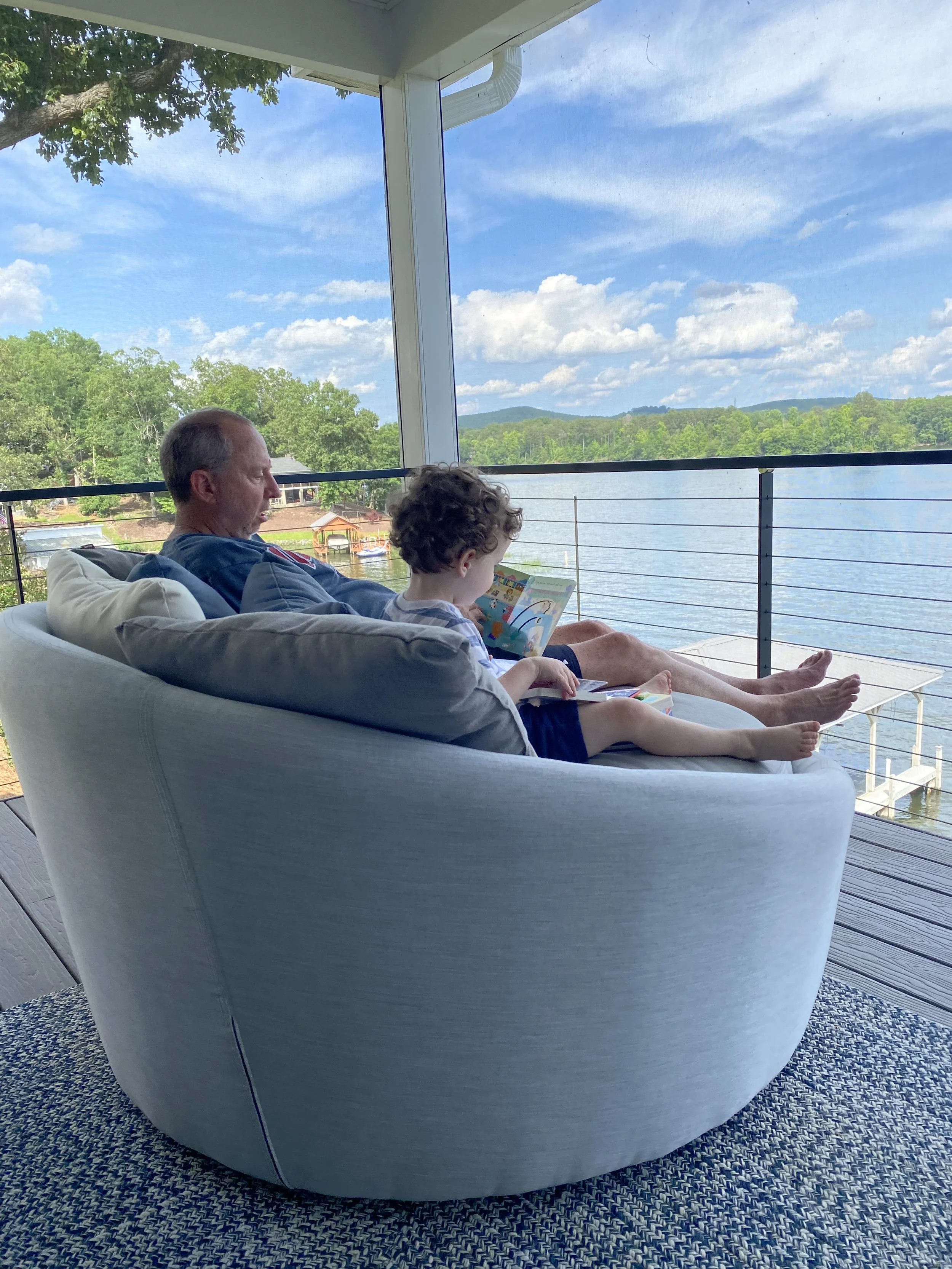 a grandpa and his grandson sitting on a screened in porch reading a story book together by the lake