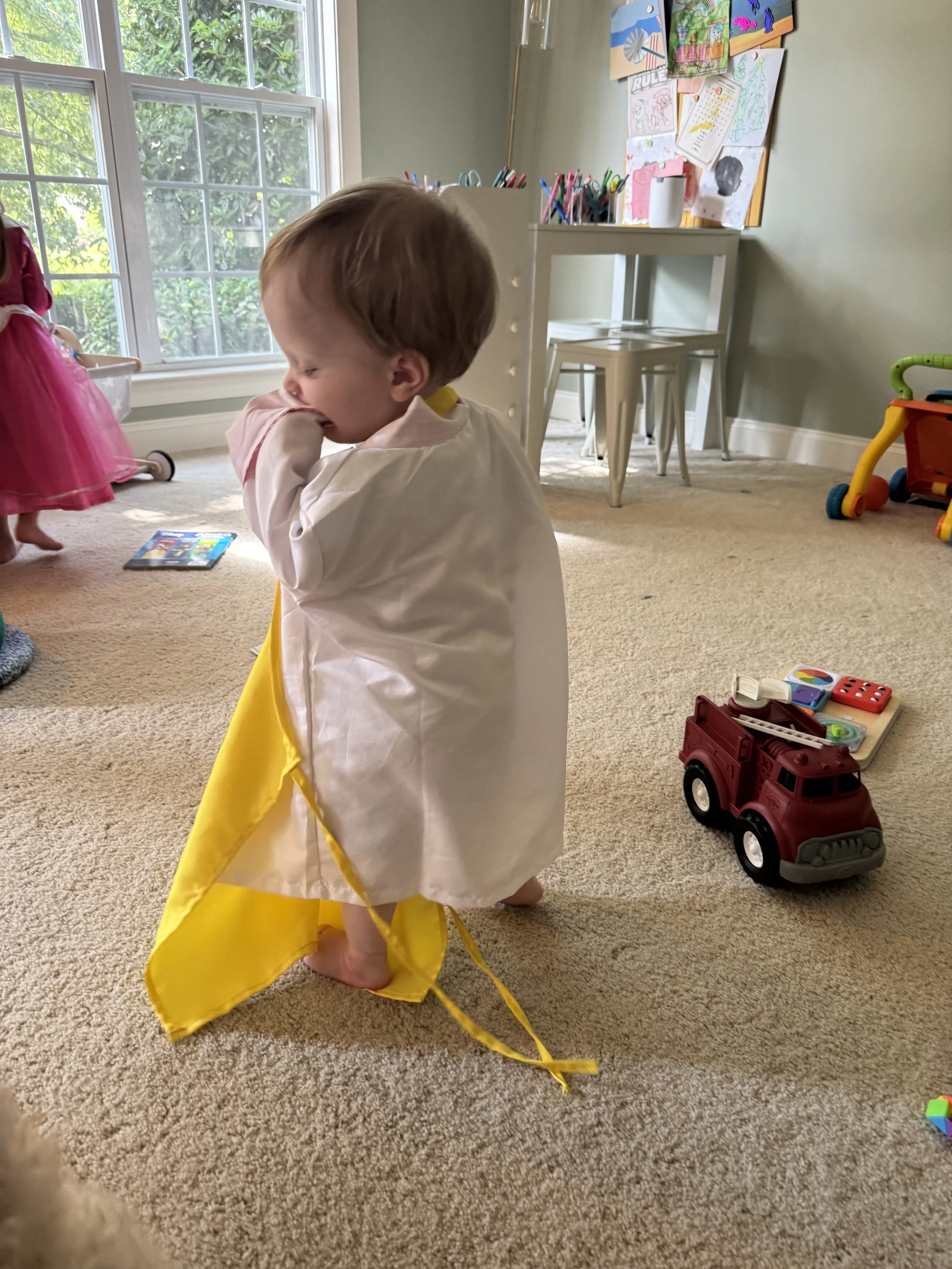a little boy in a playroom with a lab coat on and carrying a yellow apron.  he is surrounded by toys on the ground