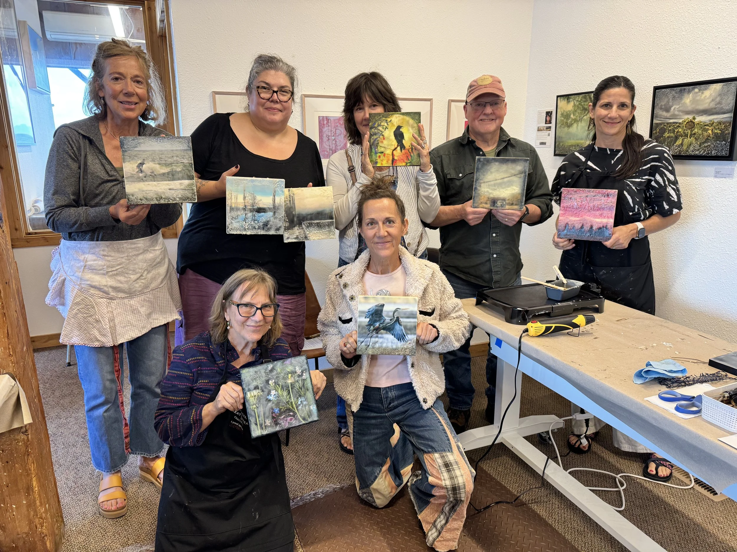 Group of eight women gathered in an art studio, holding small landscape, nature, and wildlife paintings they created, with some additional artworks hanging on the wall behind them.