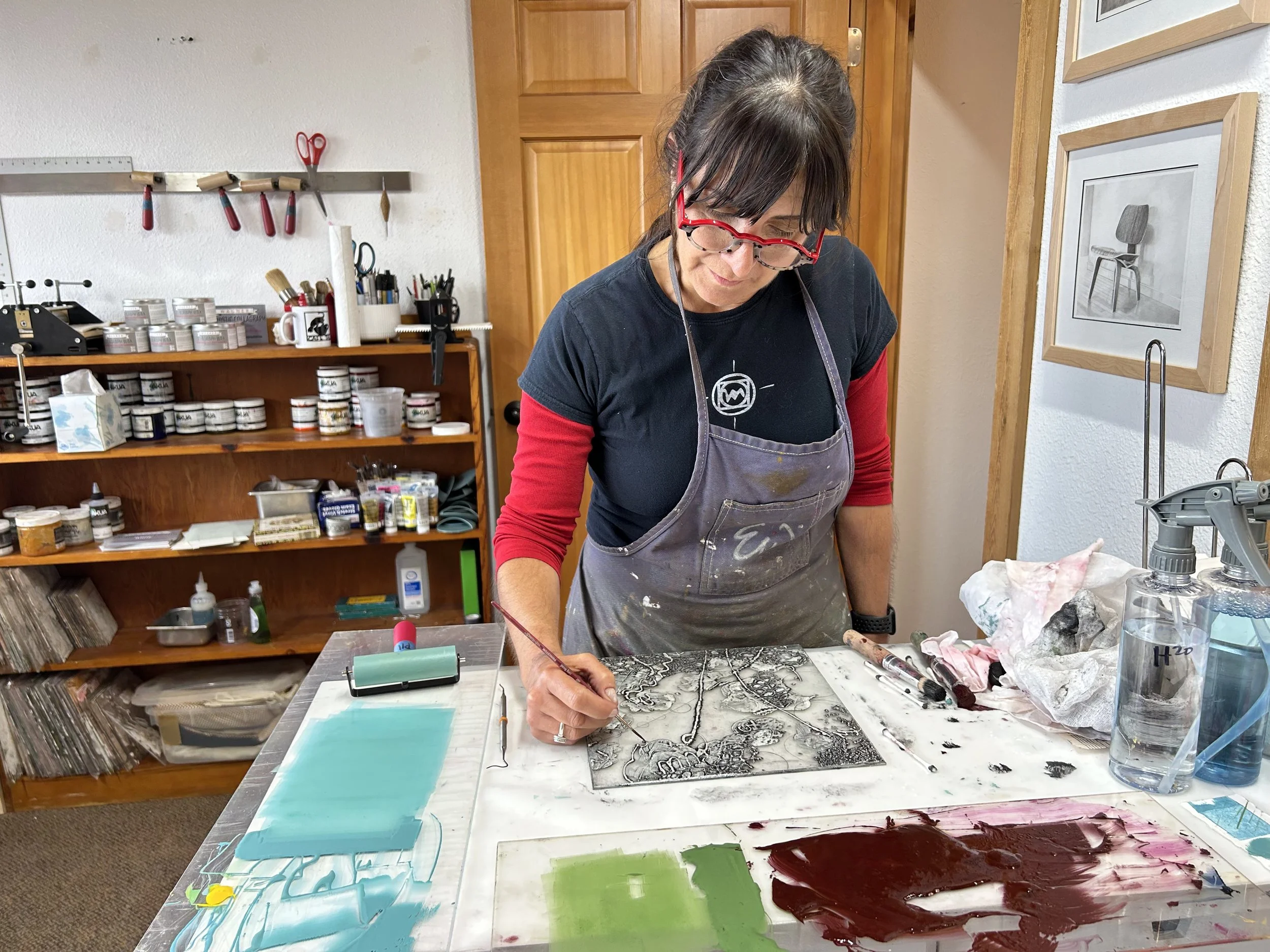 A woman wearing glasses, a black t-shirt with a logo, and an apron is working on an art piece at a table in an art studio. She is drawing or etching on a glass or metal plate while surrounded by art supplies and paints.