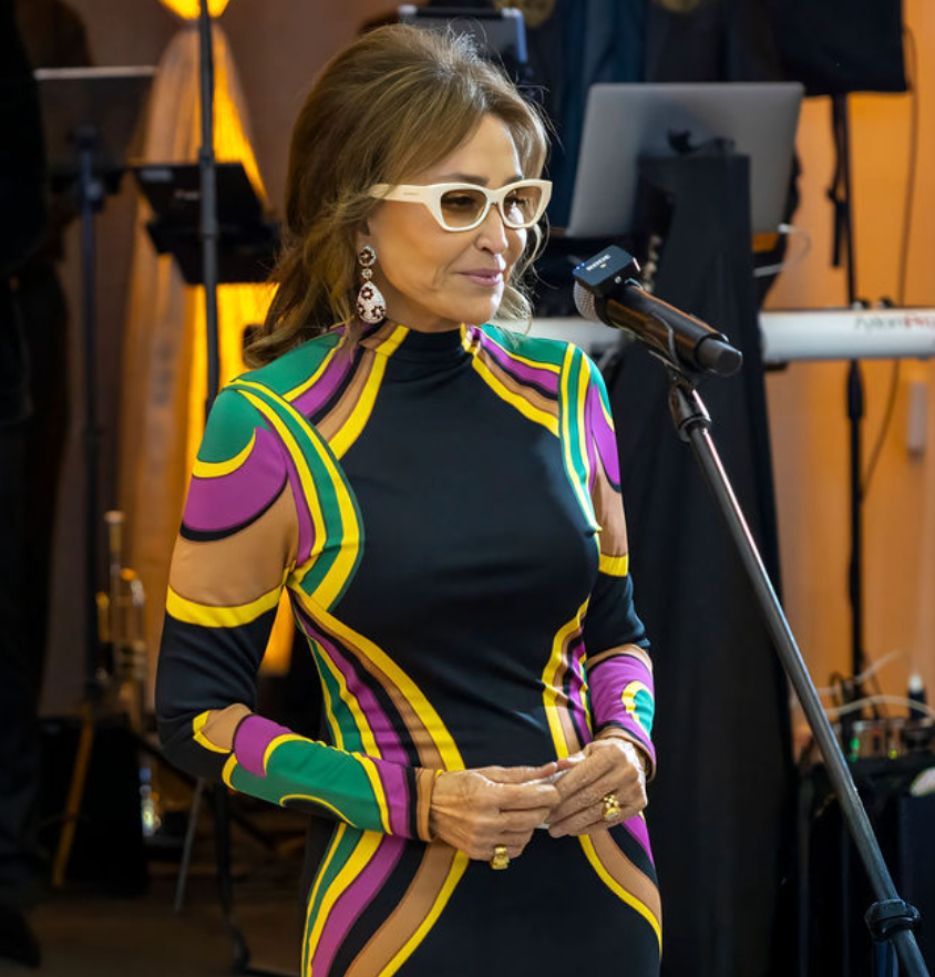A woman wearing white sunglasses, colorful patterned dress, and earrings, standing at a microphone in a room with musical equipment.
