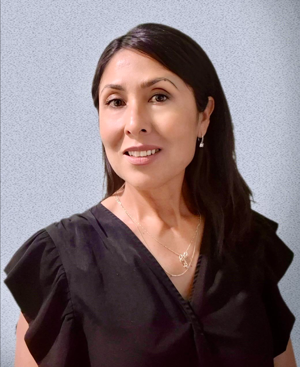 A woman with dark hair smiling, wearing earrings and layered necklaces, in front of a light gray textured background.
