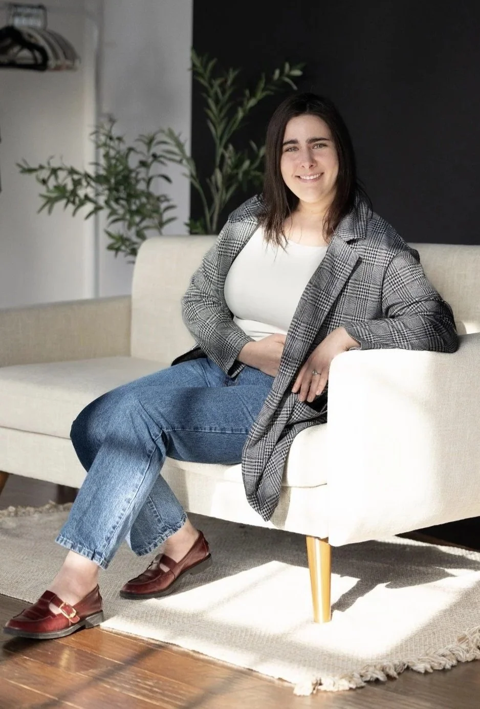 A woman with brown hair sitting on a beige couch in a modern living room, smiling, wearing a plaid blazer, white top, blue jeans, and red loafers, with green plants in the background.