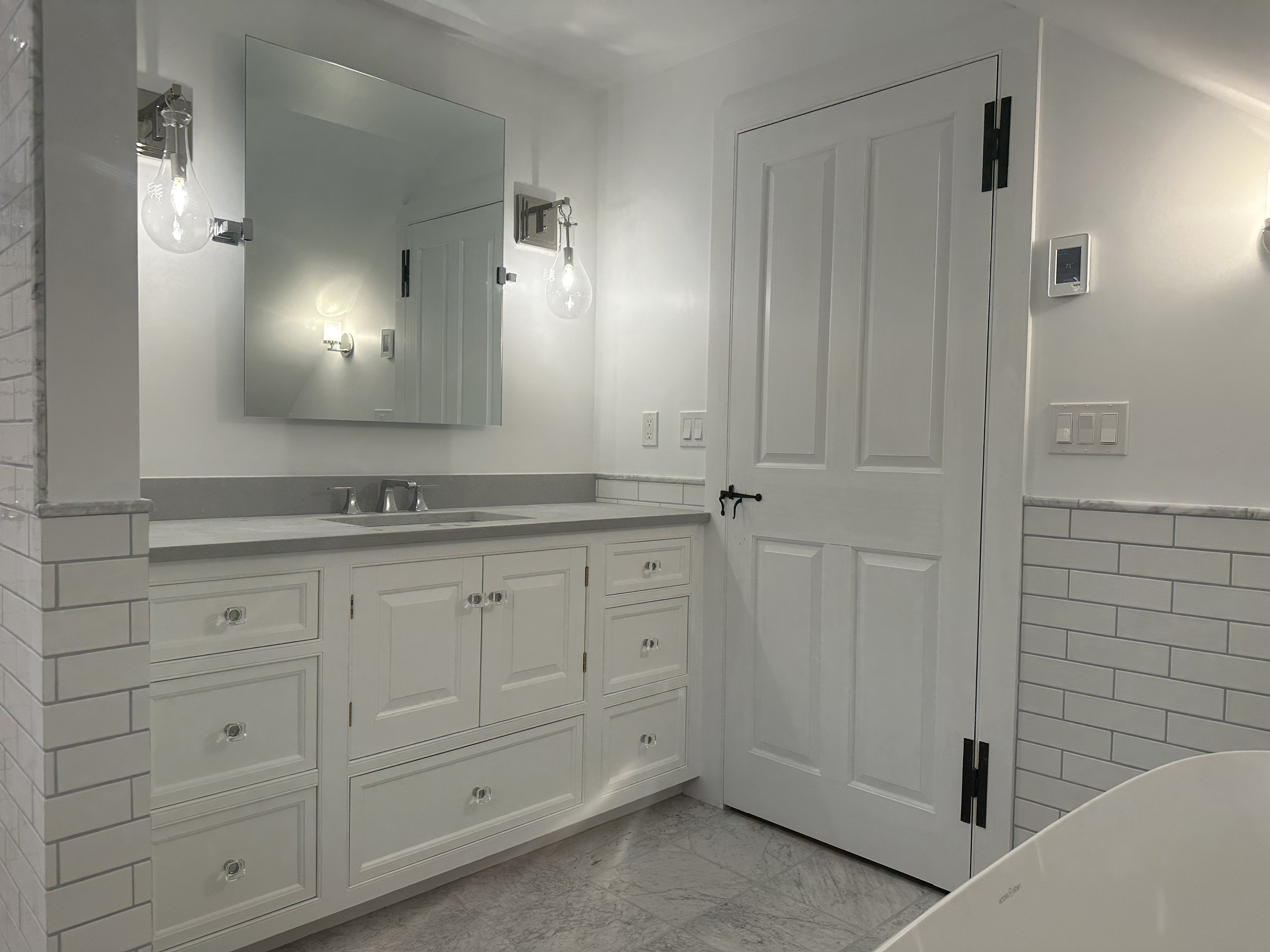 A modern bathroom with white cabinetry and a gray countertop, a wall-mounted mirror, and two pendant lights with exposed bulbs. There is a door with black hinges and a black latch, white subway tile wainscoting, and a thermostat on the wall.