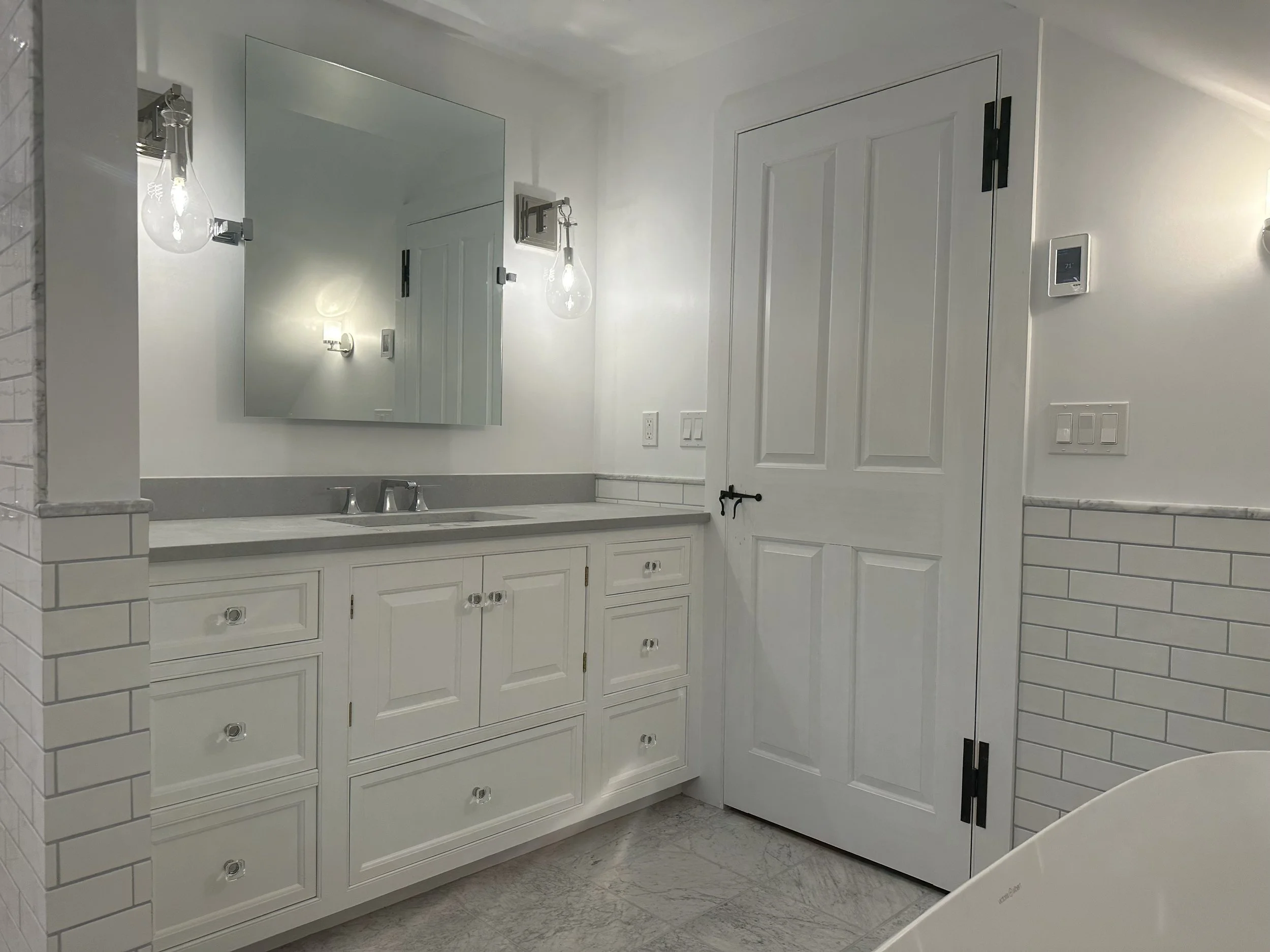 A bright bathroom with white cabinetry, a large mirror, and modern lighting fixtures, featuring a white door and tiled walls.