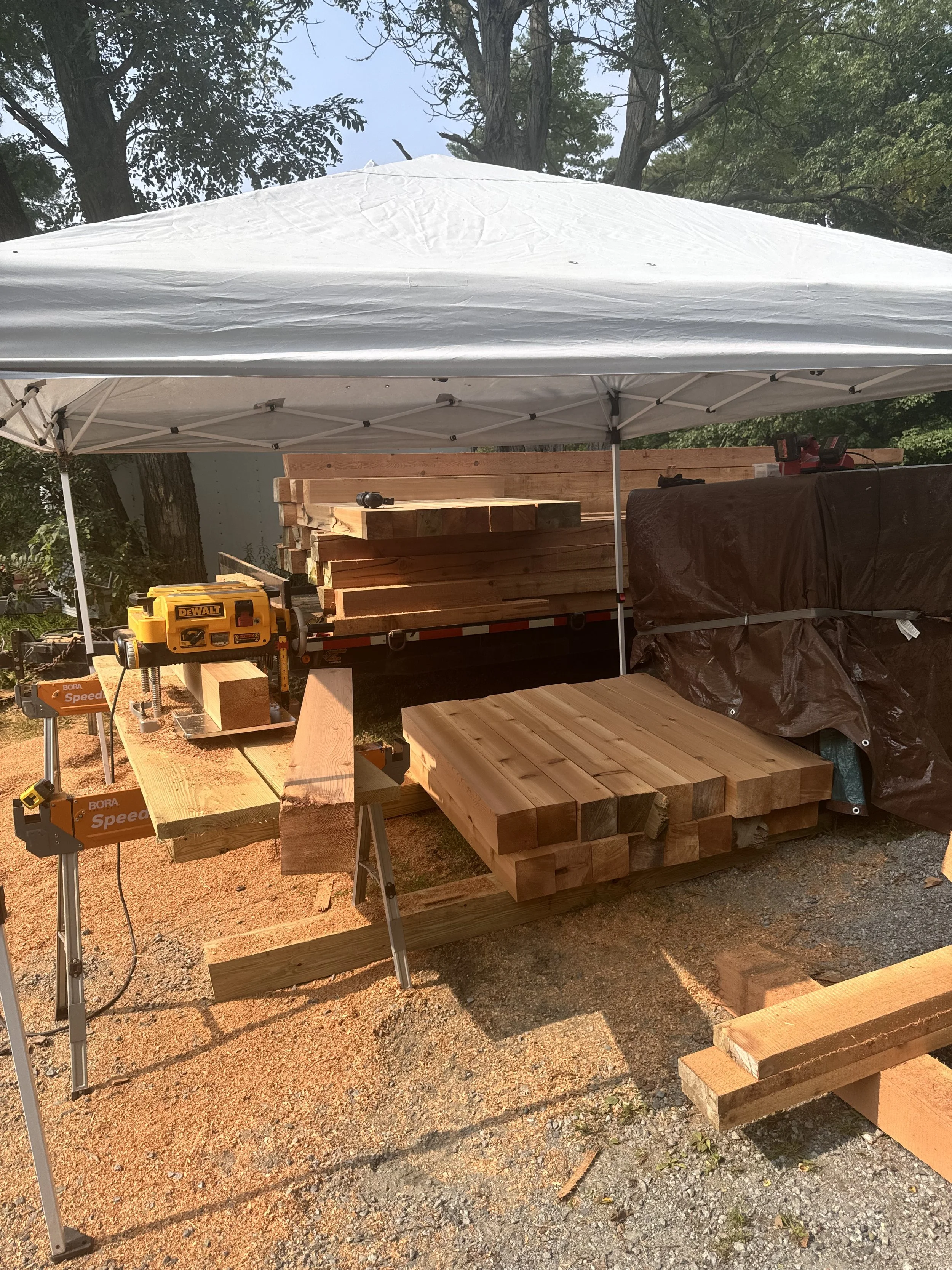 Woodworking setup with stacked lumber, a workbench, and power tools under a white canopy outdoors.