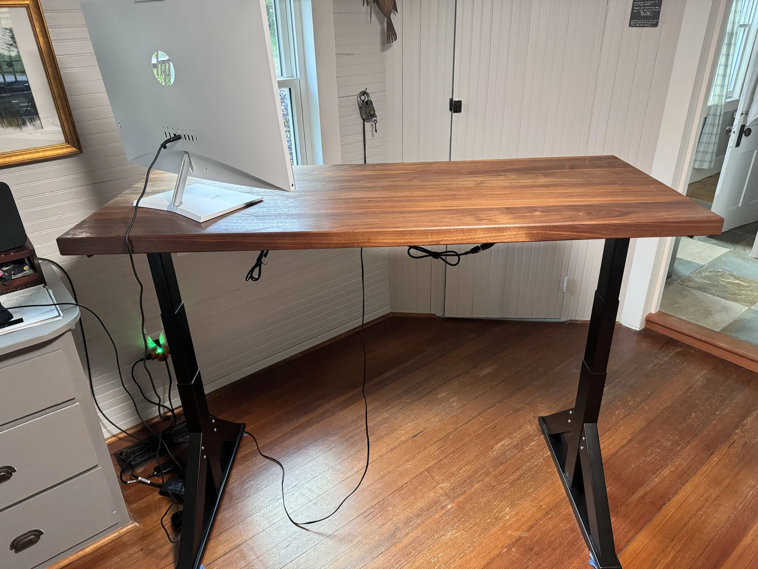 A standing desk with a large white monitor, wires hanging from the back, situated in a room with wood flooring and white walls.