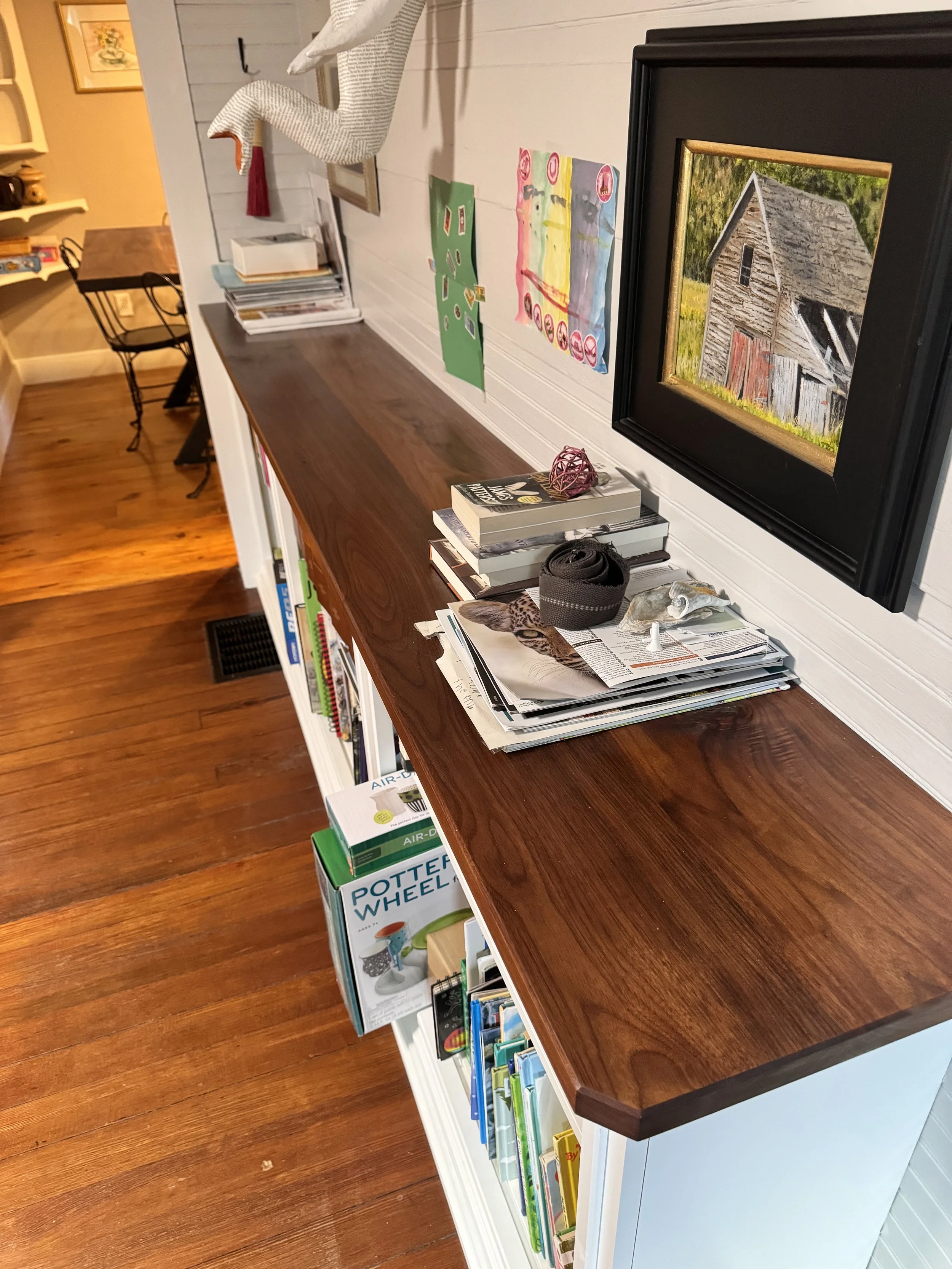A wooden sideboard or bookshelf with books, magazines, and decorative items on top, located in a room with wooden flooring and white walls decorated with artwork.