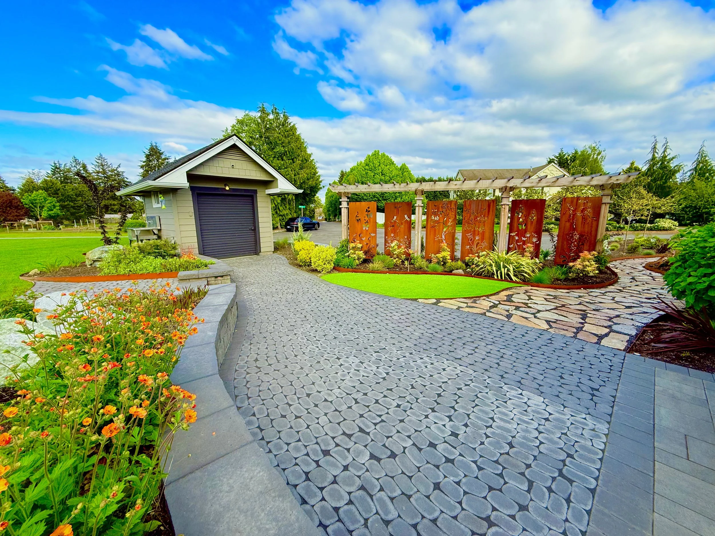 Colorful landscaped backyard with paved stone walkway, garden beds, and a small shed under a blue sky with scattered clouds.