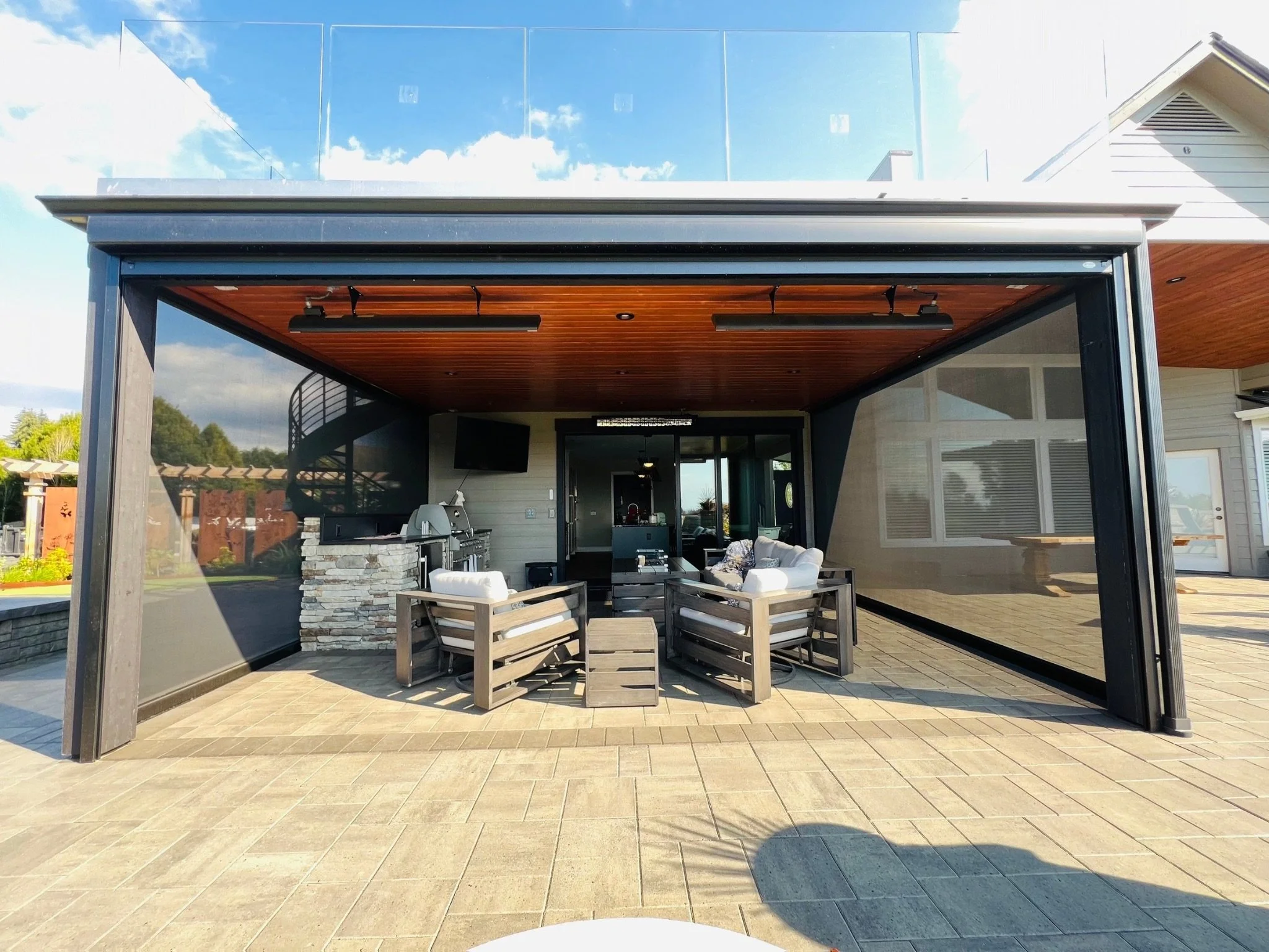 Outdoor patio area with seating furniture, a grill, and a large glass-enclosed space in the background, with a partly cloudy sky above.