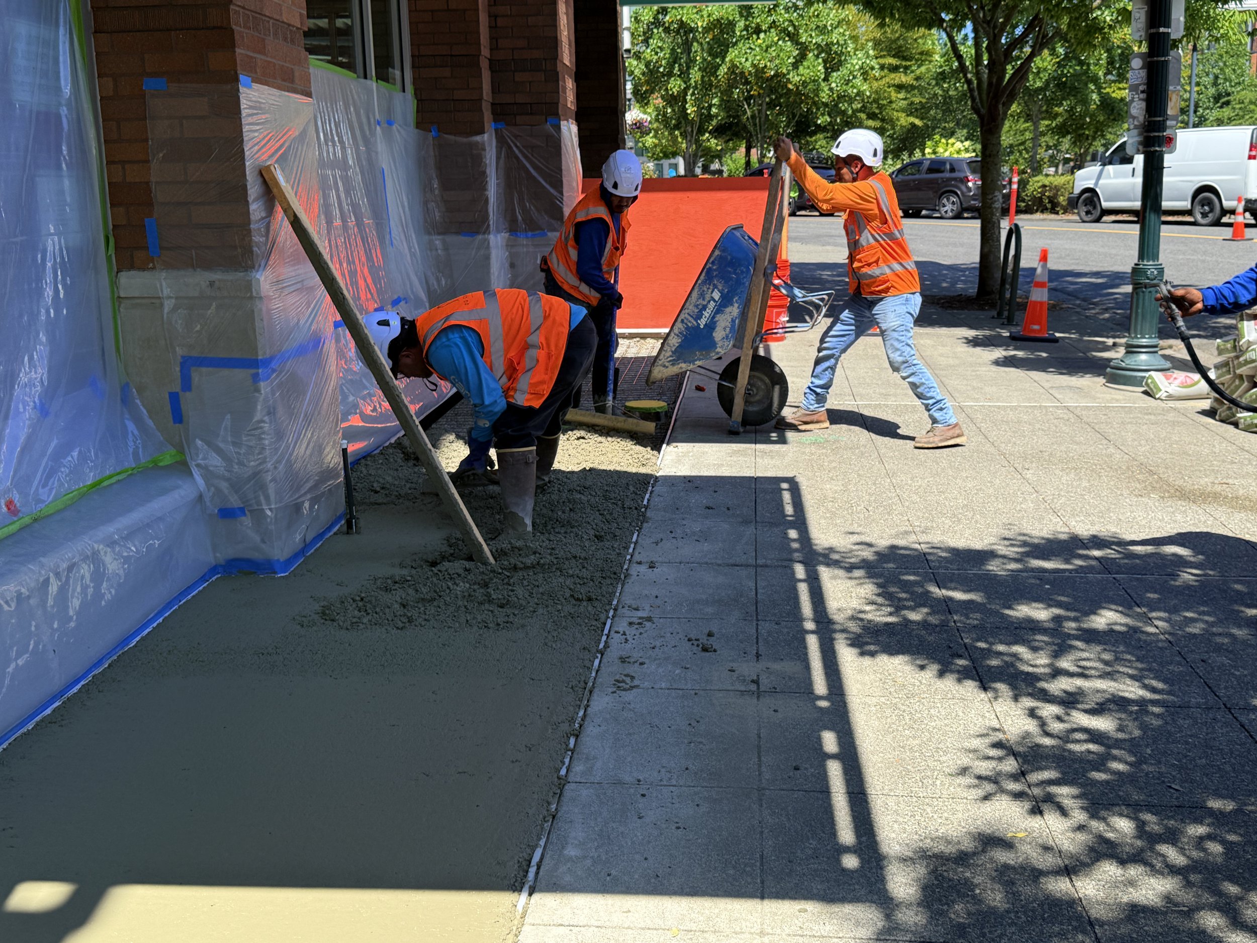 Construction workers in orange safety vests and helmets pouring and leveling concrete on a sidewalk. One worker shovels wet concrete, another uses a hand float, and others are finishing the surface.