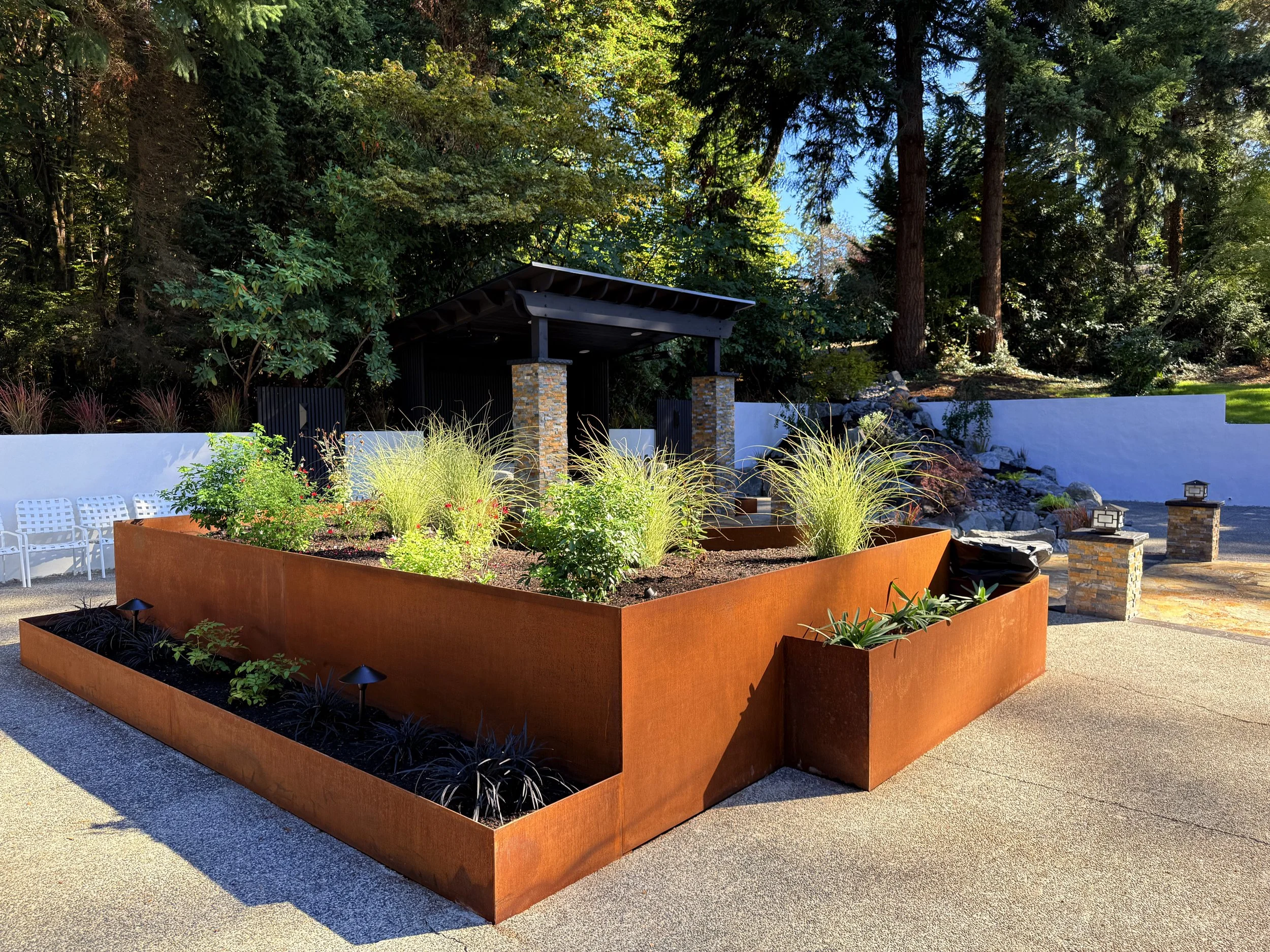 Modern outdoor garden with rust-colored raised planters filled with plants, trees, and grasses, surrounded by a paved patio, with a white wall, benches, and a small shed in the background, and large trees providing shade.