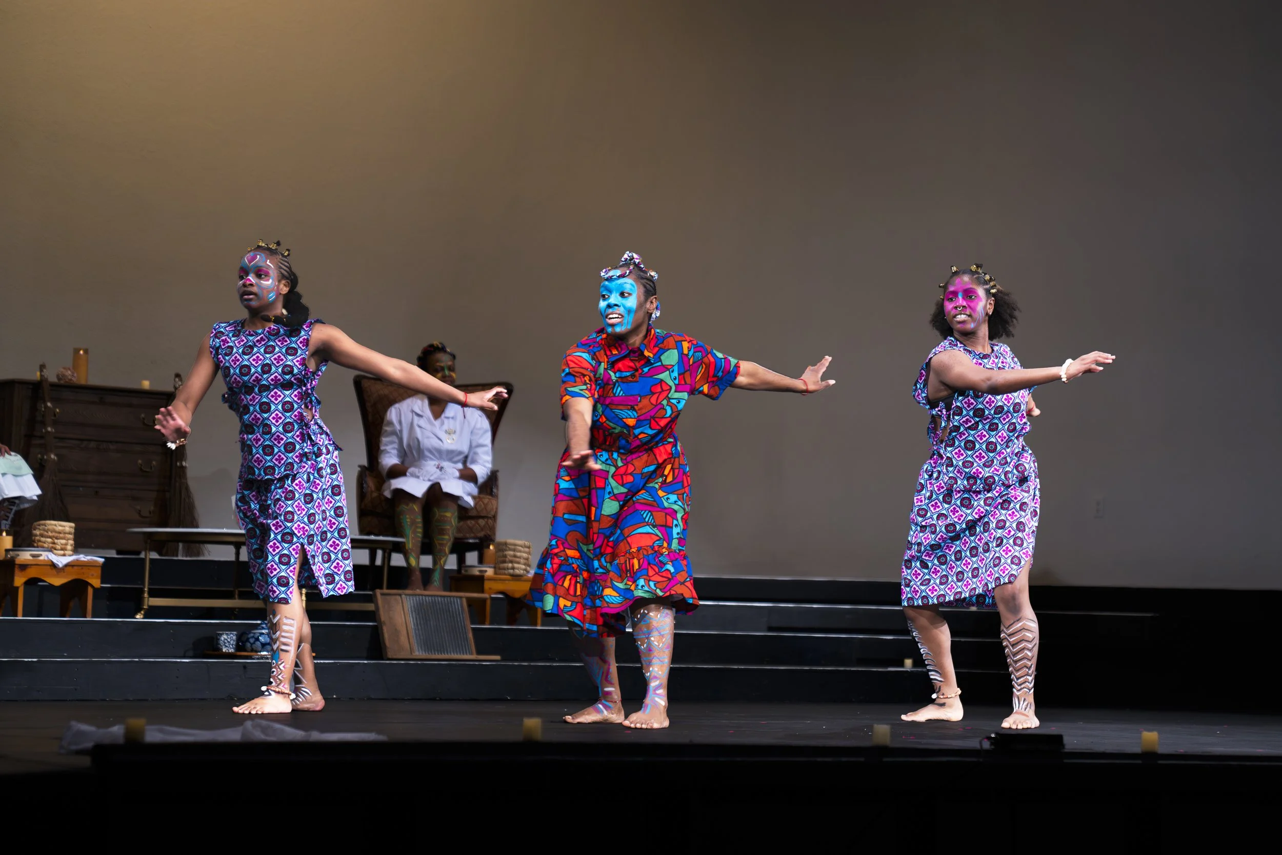 Three children with colorful face paint performing a dance on stage, with an adult seated in the background.