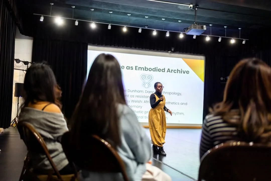 A woman in a yellow jumpsuit speaking at a presentation with an audience seated in front of her in a dark room with a large screen behind her.