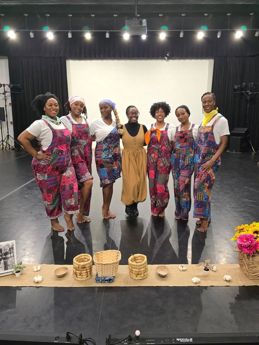 A group of seven women standing on a stage, smiling at the camera, wearing colorful, patterned overalls and white t-shirts. No shoes, some with head scarves. In front of them is a wooden table with baskets, flowers, and decorative items. The stage has black curtains and stage lights overhead.