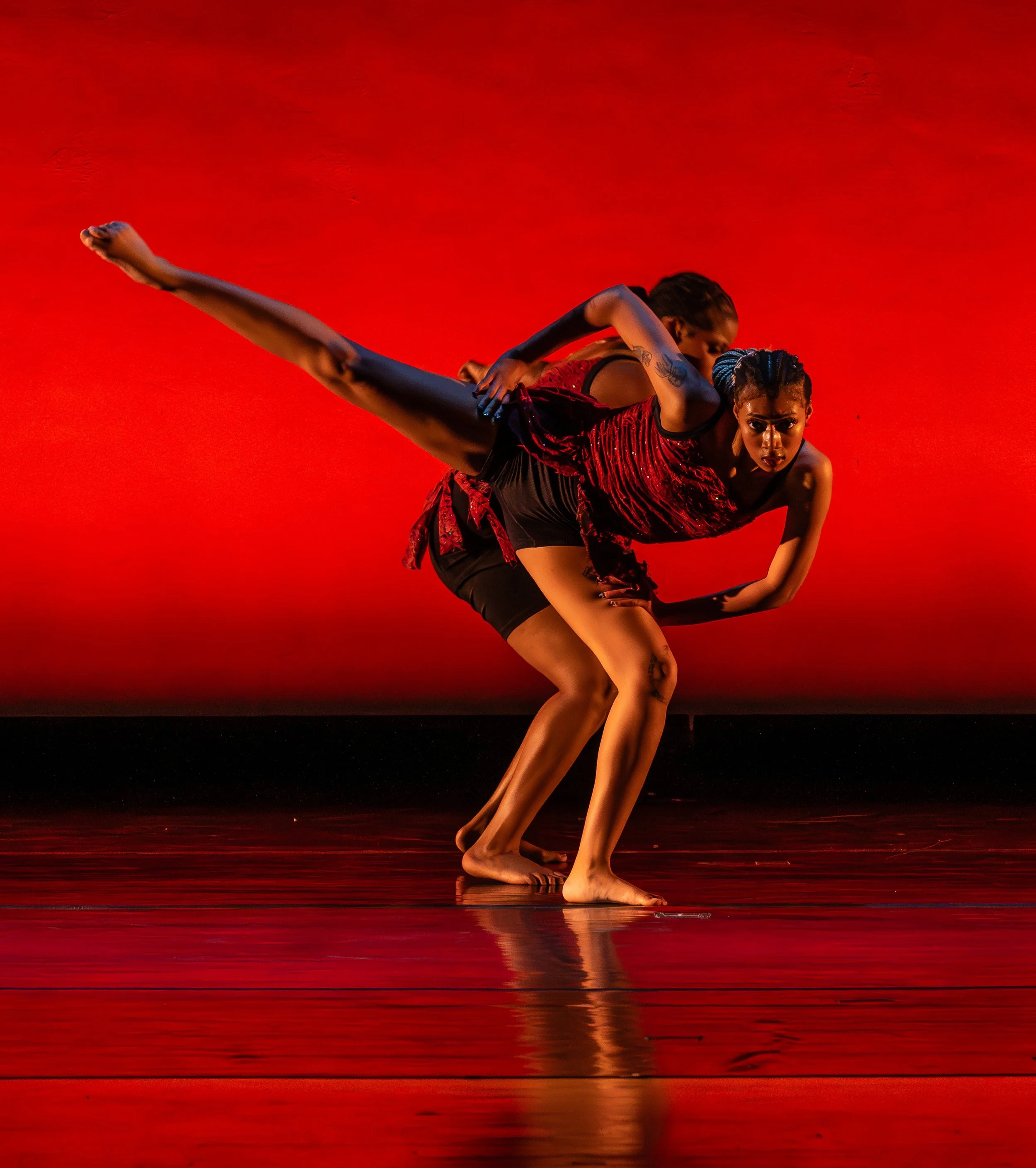Two dancers performing a contemporary dance move on stage with a red background.