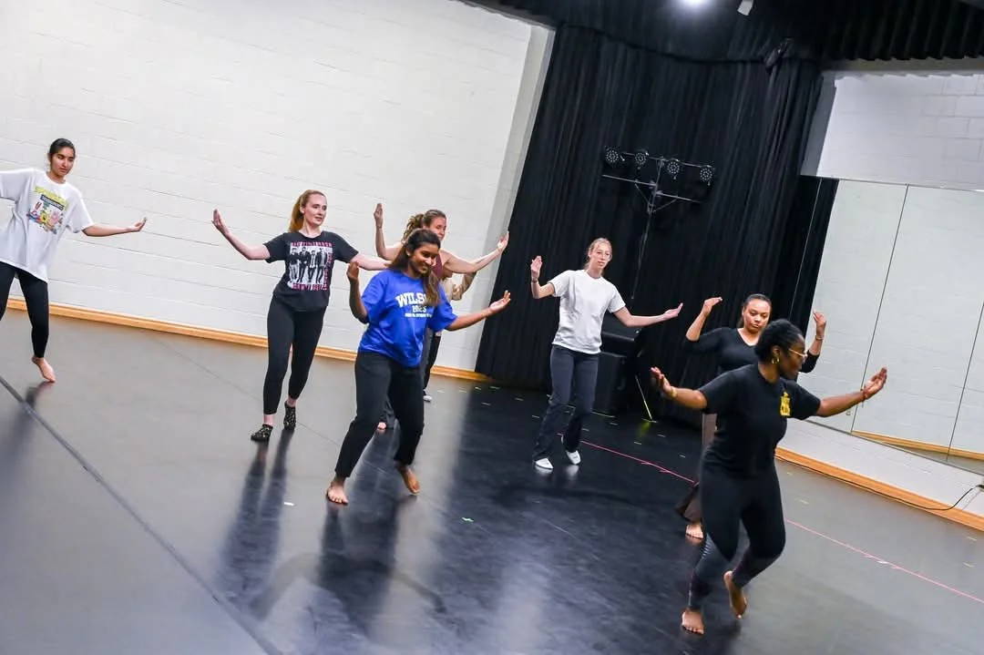 A group of women practicing dance or choreography in a dance studio with mirrors and black curtains.