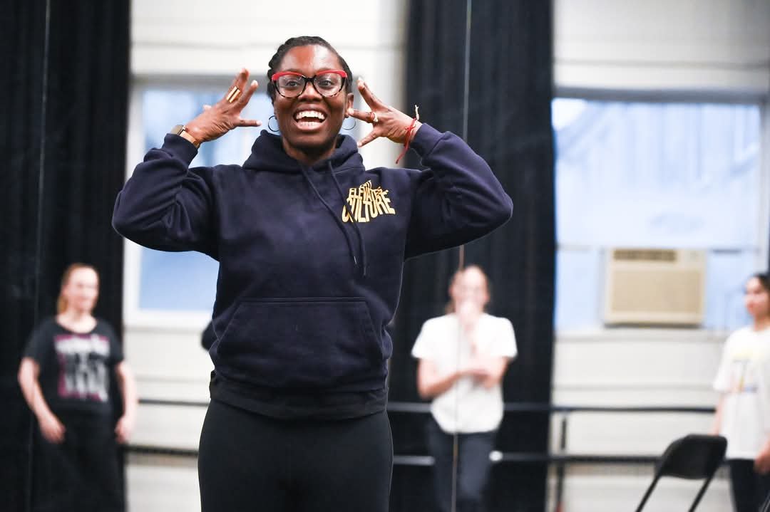 A woman with glasses, smiling, wearing a black hoodie, and standing in a dance or rehearsal studio with others in the background.