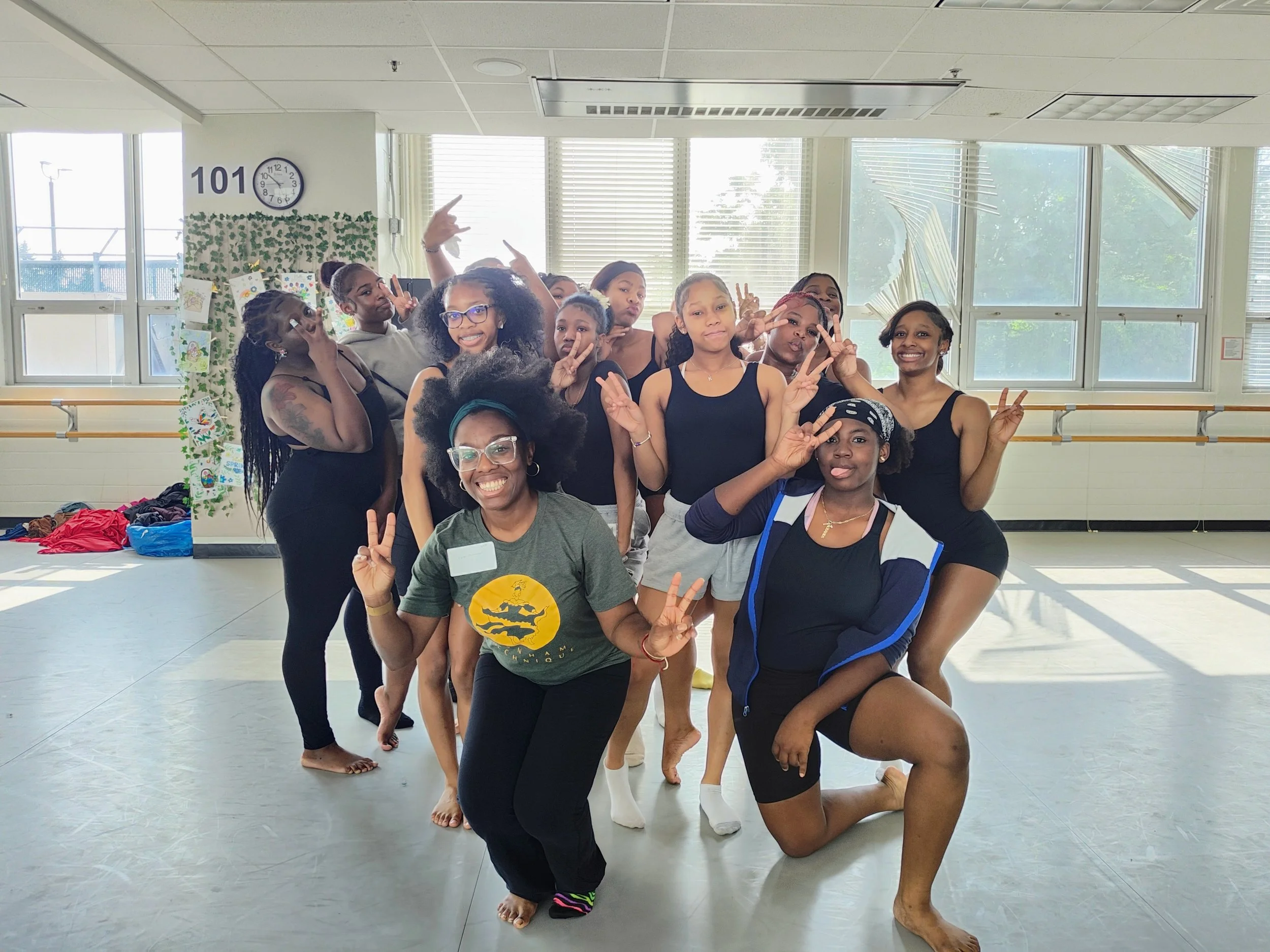 A group of diverse girls and women in workout clothes posing and making peace signs in a dance studio with large windows, a clock, and a decorative wall.