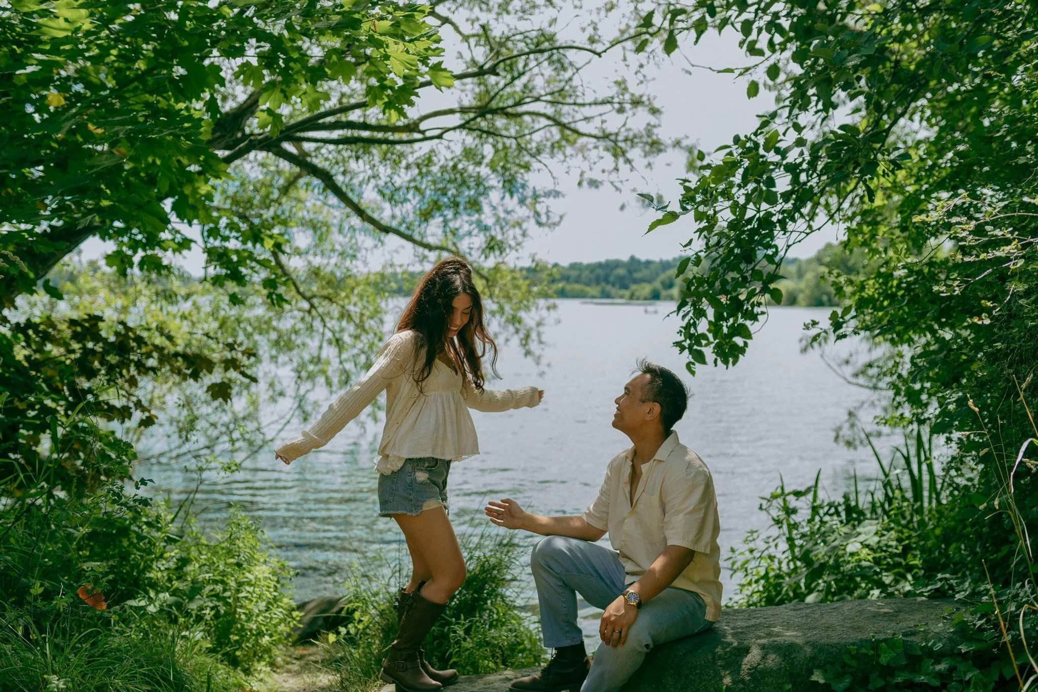 A young girl being lifted by a man near a lake, surrounded by green trees and foliage on a sunny day.